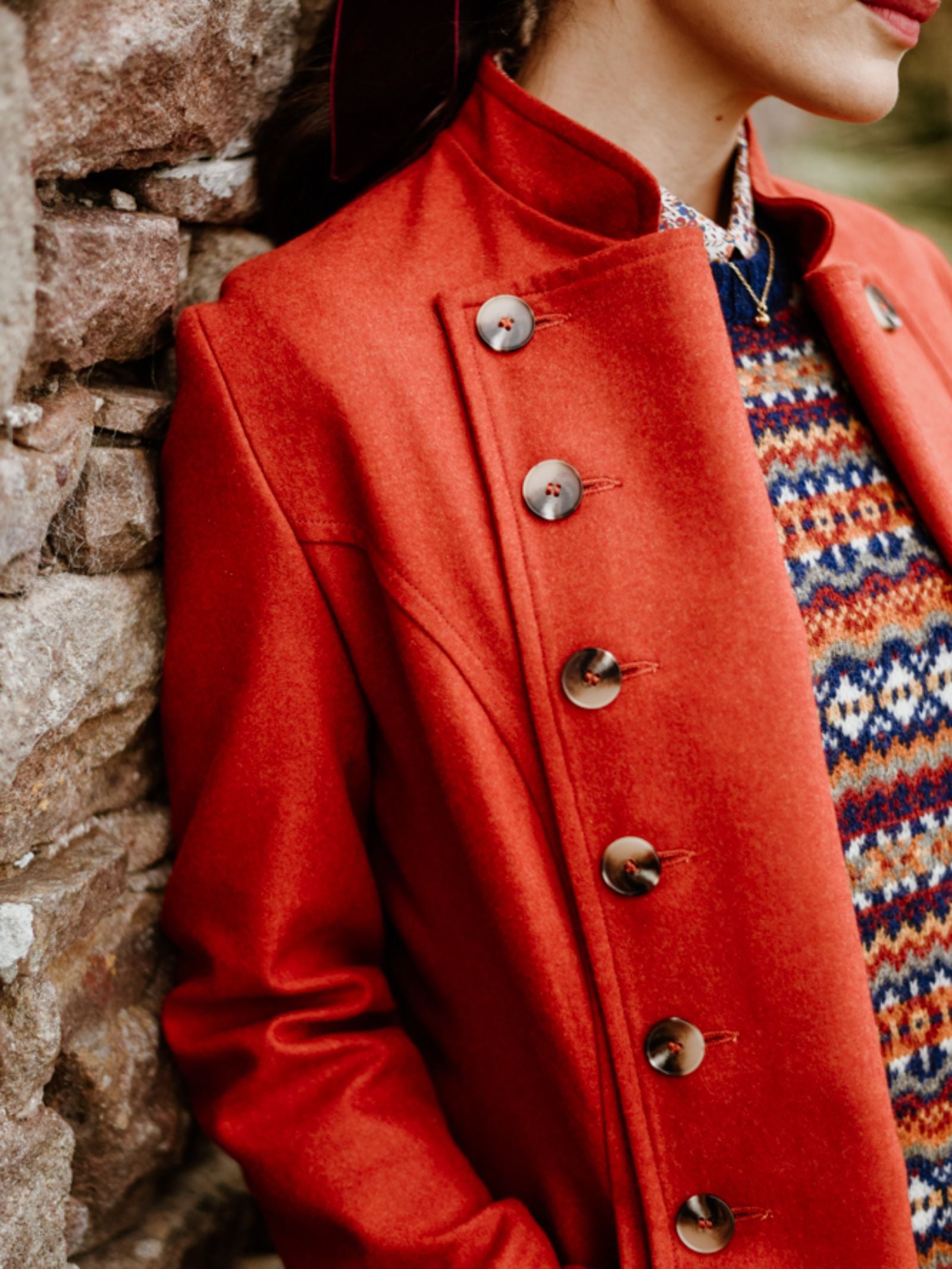 A person wearing a vibrant red Pirate Coat by Campbell's of Beauly with large buttons and a colorful patterned sweater underneath leans against a stone wall. The image is cropped to show their torso and part of their face.