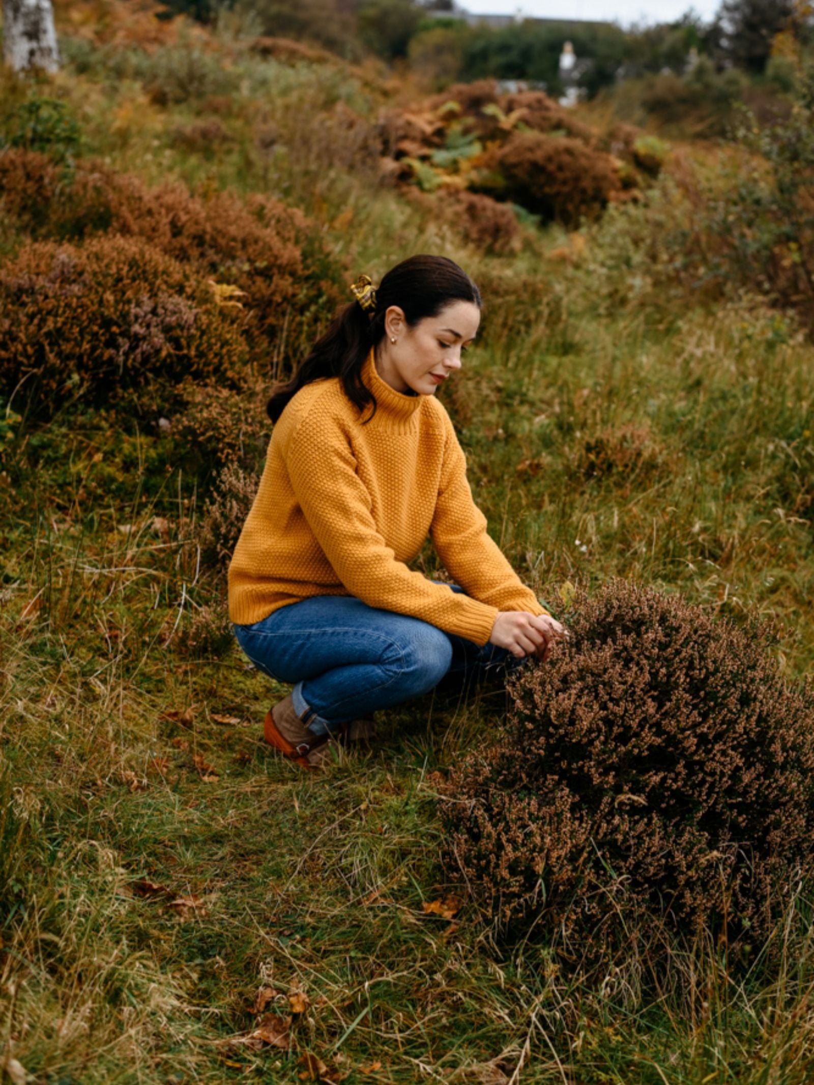 A woman in a mustard yellow Chunky Moss Stitch Polo by Campbell’s of Beauly and blue jeans crouches outdoors, touching bush leaves amid grass and autumn foliage.