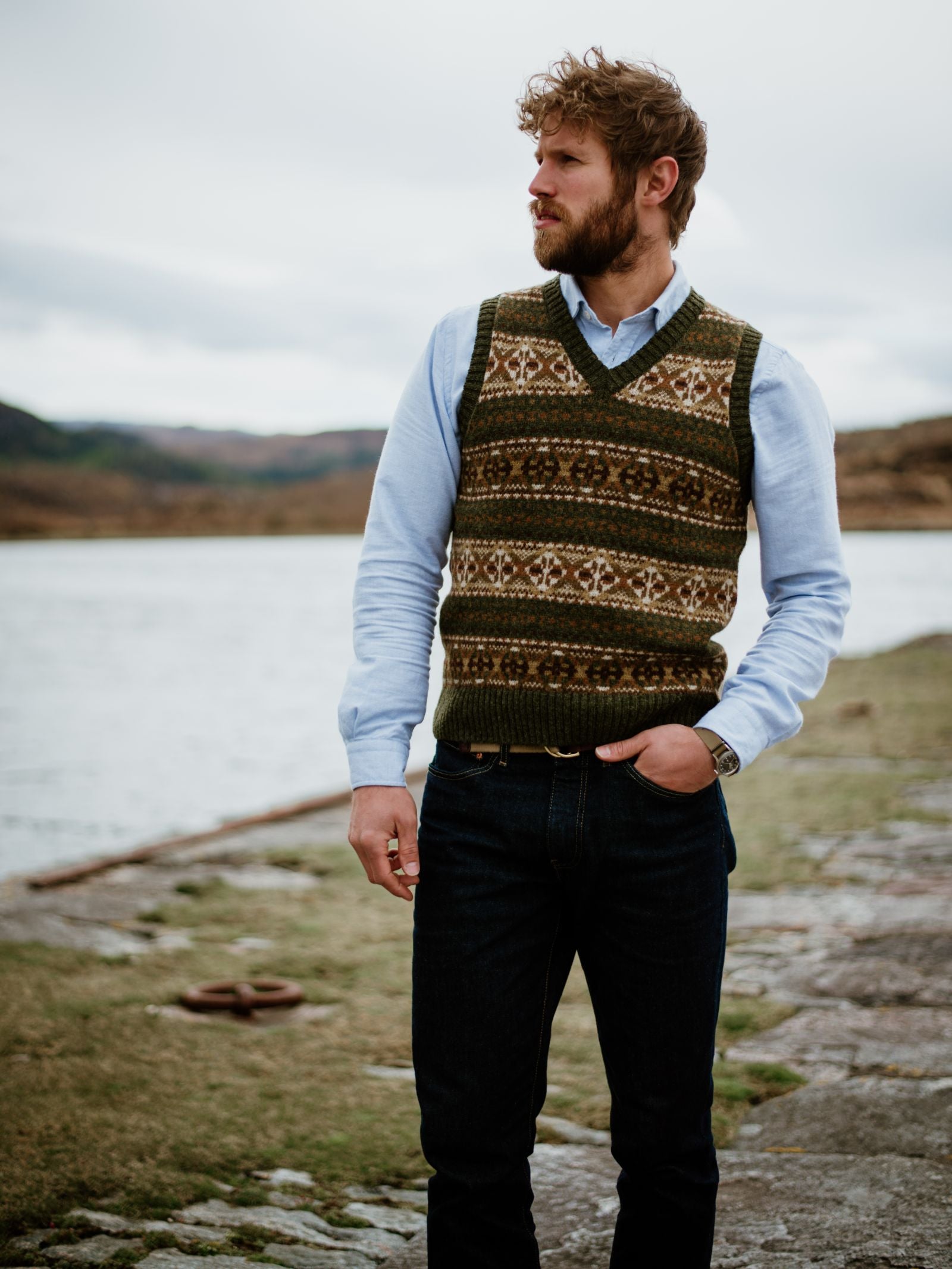 A bearded man with curly brown hair stands by a lake in a blue shirt, dark jeans, and the Campbells of Beauly Fairisle Slipover, a sleeveless Shetland wool knit. He looks to the side with one hand in his pocket.