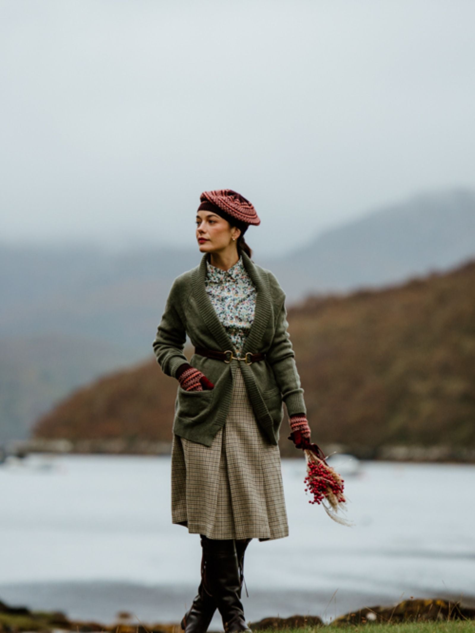 A woman stands by a lake, holding a bouquet. She wears a Campbell's of Beauly Cashmere Shawl Cardigan, patterned beret, floral blouse, plaid skirt, and tall boots. Misty hills form a dreamy outdoor backdrop.