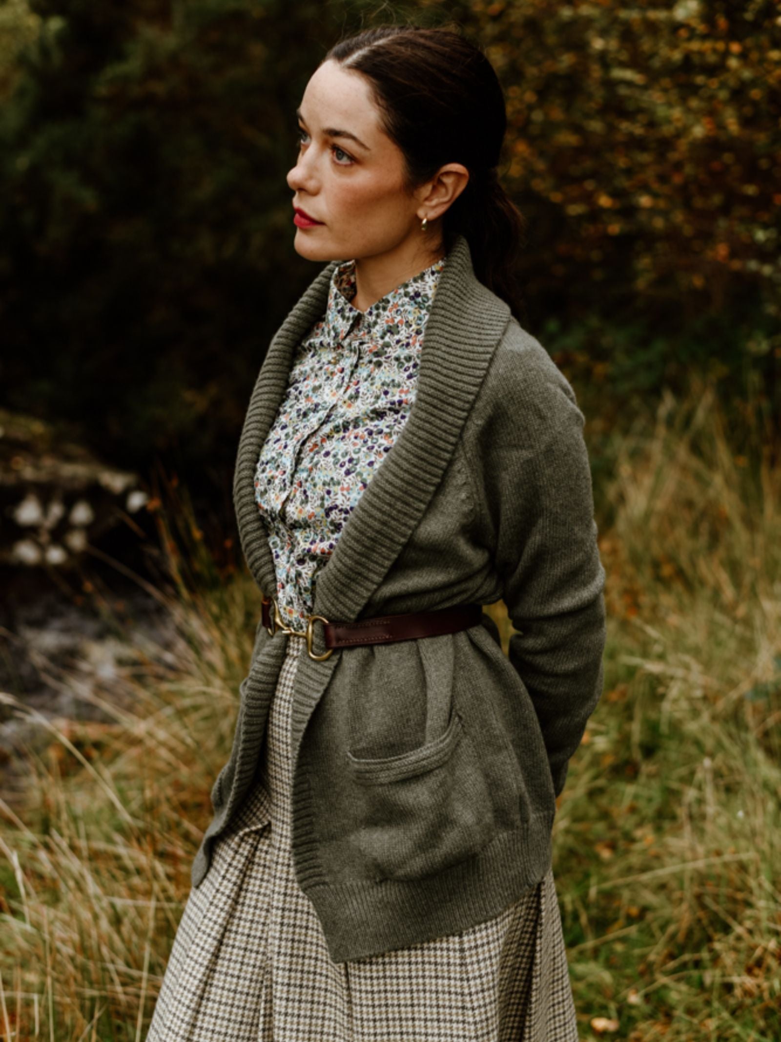 A woman stands in tall grass, wearing a Campbell's of Beauly Cashmere Shawl Cardigan belted at the waist over a floral blouse and plaid skirt, looking thoughtfully to the side with autumn trees behind her.