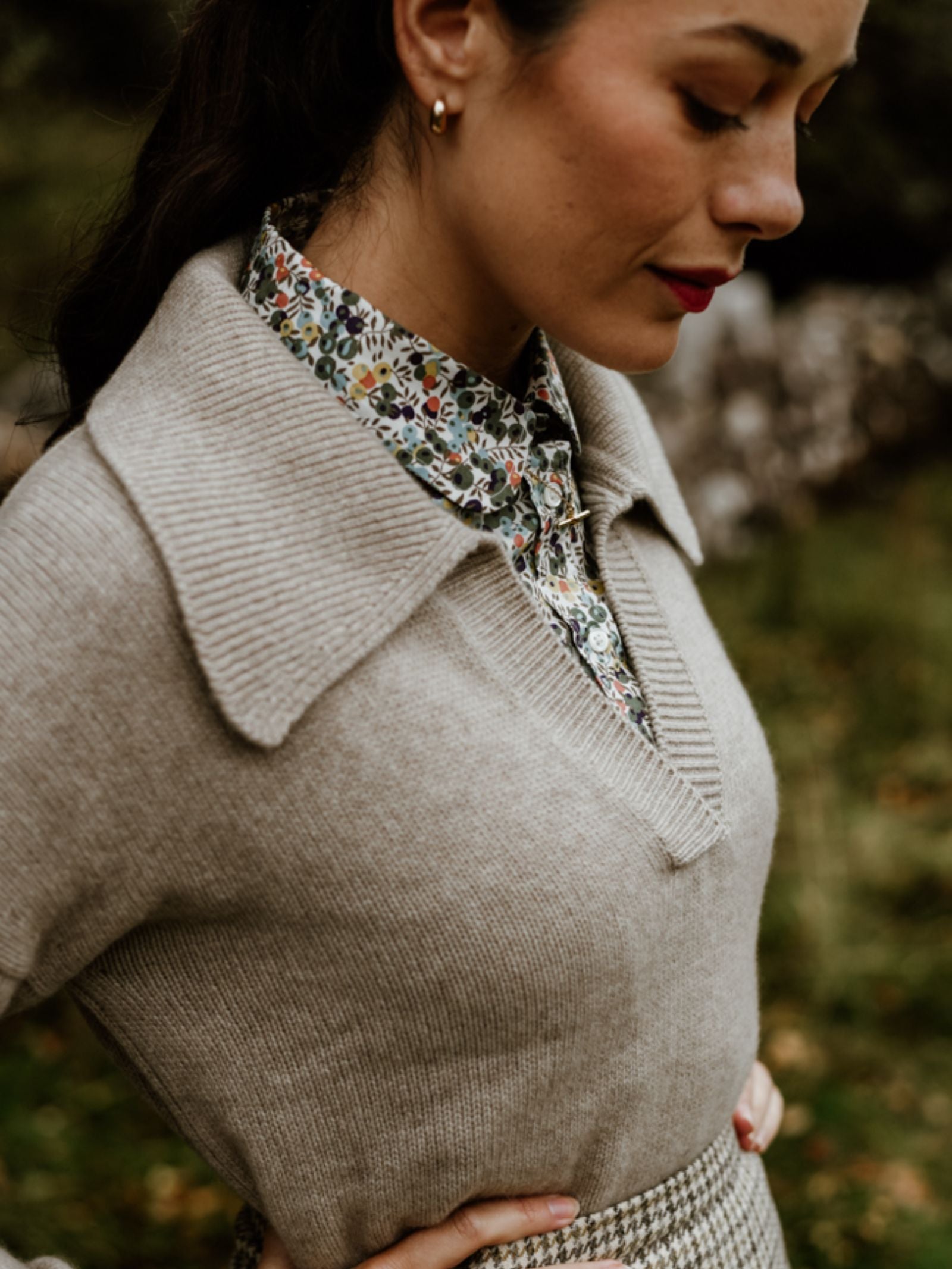 A woman stands outdoors in the Campbell's of Beauly Cashmere Shirt Jumper layered over a floral collared shirt. Her dark hair is tied back, gold hoop earrings shine, and she gazes down gently amid blurred greenery.