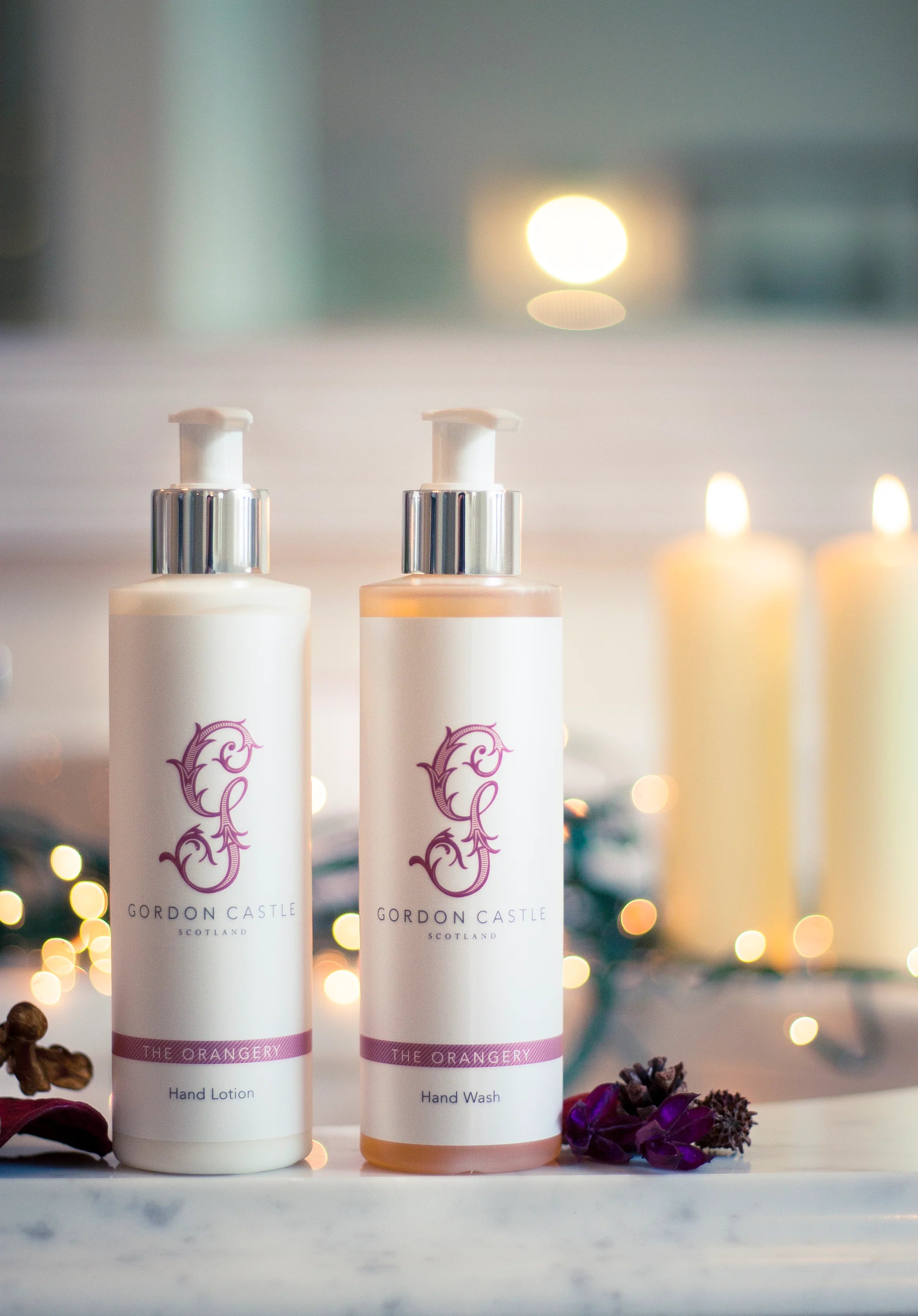 Two bottles—Gordon Castle Orangery Hand Lotion and Hand Wash with sweet orange and mandarin oils—rest on a marble surface, while blurred candles and string lights create a cozy background.