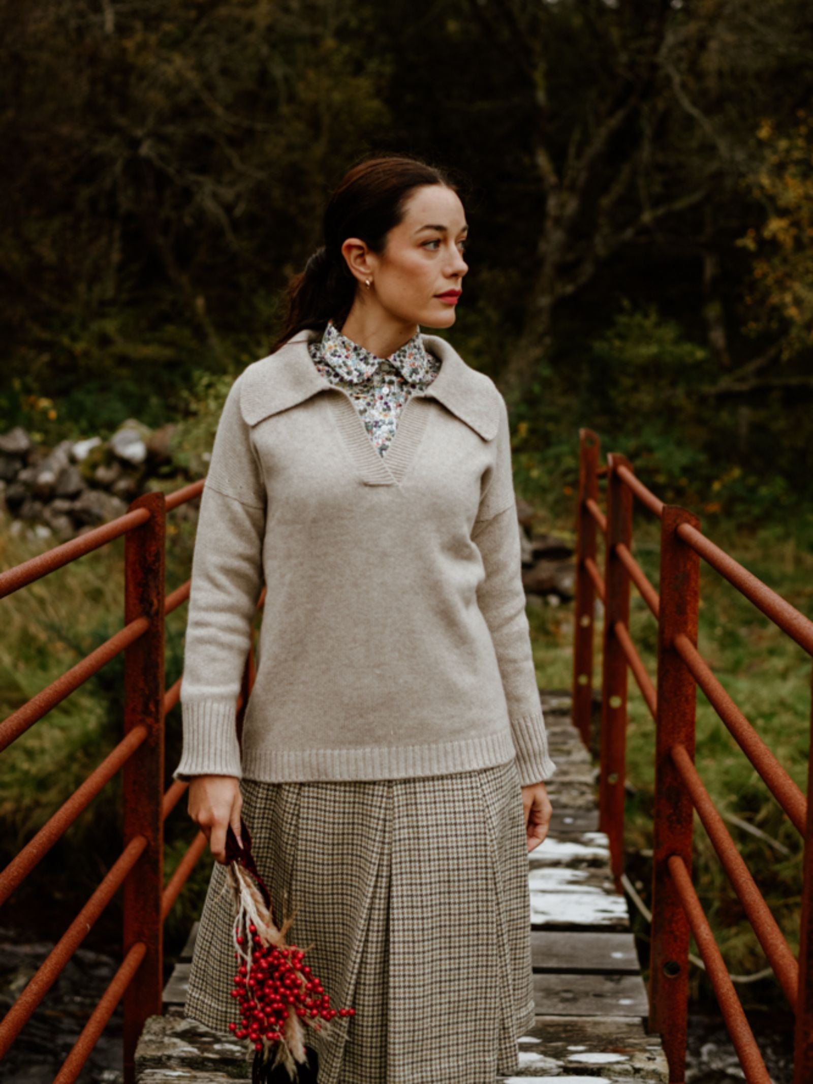 A woman in the Campbell's of Beauly Cashmere Shirt Jumper and a plaid skirt stands on a rustic wooden bridge with red railings, holding red berries amid lush trees and greenery inspired by the Scottish Borders.