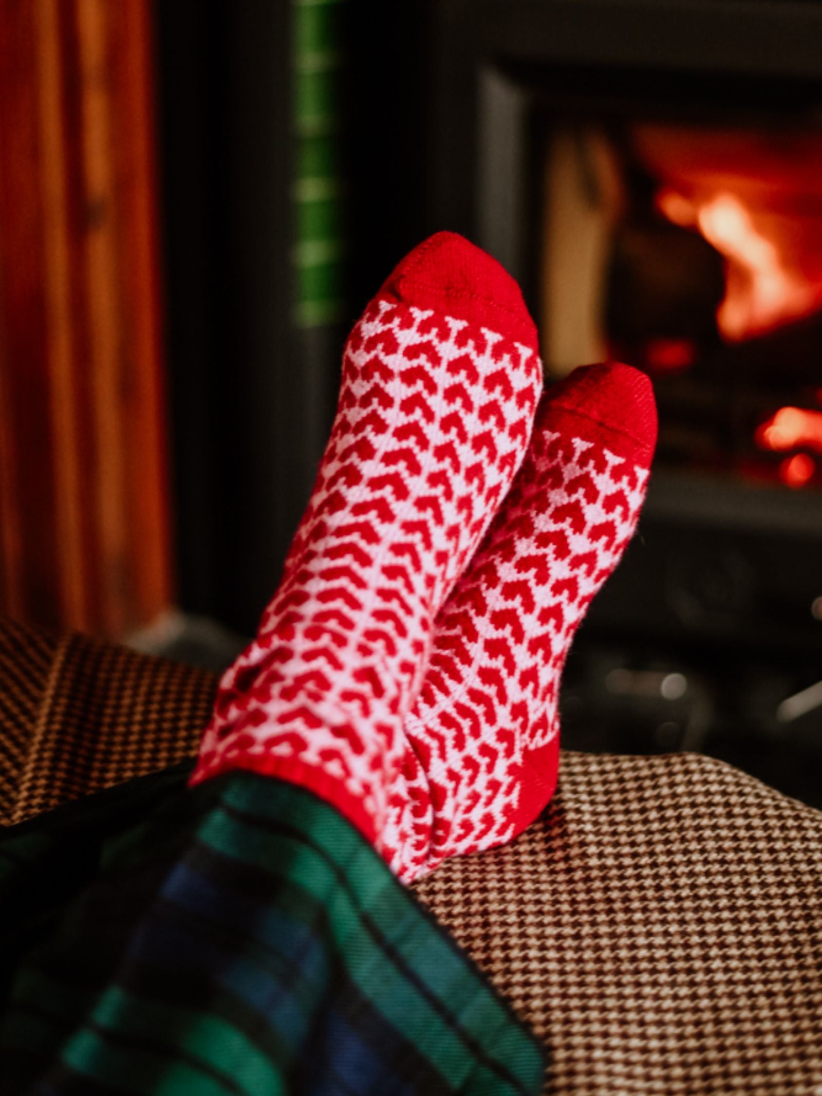 Someone wearing Campbell's of Beauly Heart Socks Red, featuring a red and white heart pattern, relaxes by a fireplace in plaid pajama pants, creating a warm and cozy atmosphere.