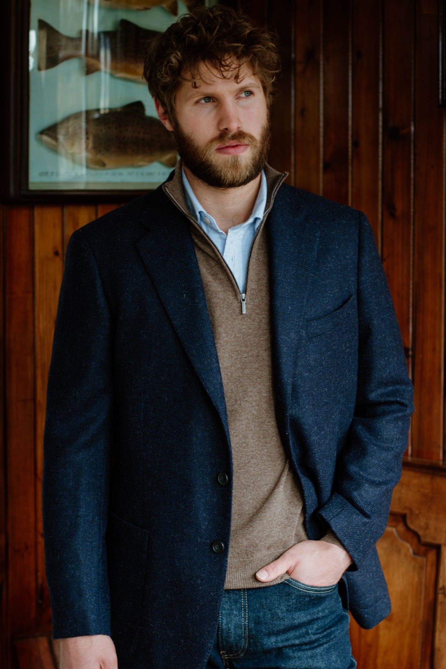 A bearded man with curly hair wears a Campbells of Beauly Merino & Cashmere Quarter Zip Jumper under a navy blazer and light blue shirt, standing indoors against wooden paneling with framed fish artwork behind him.
