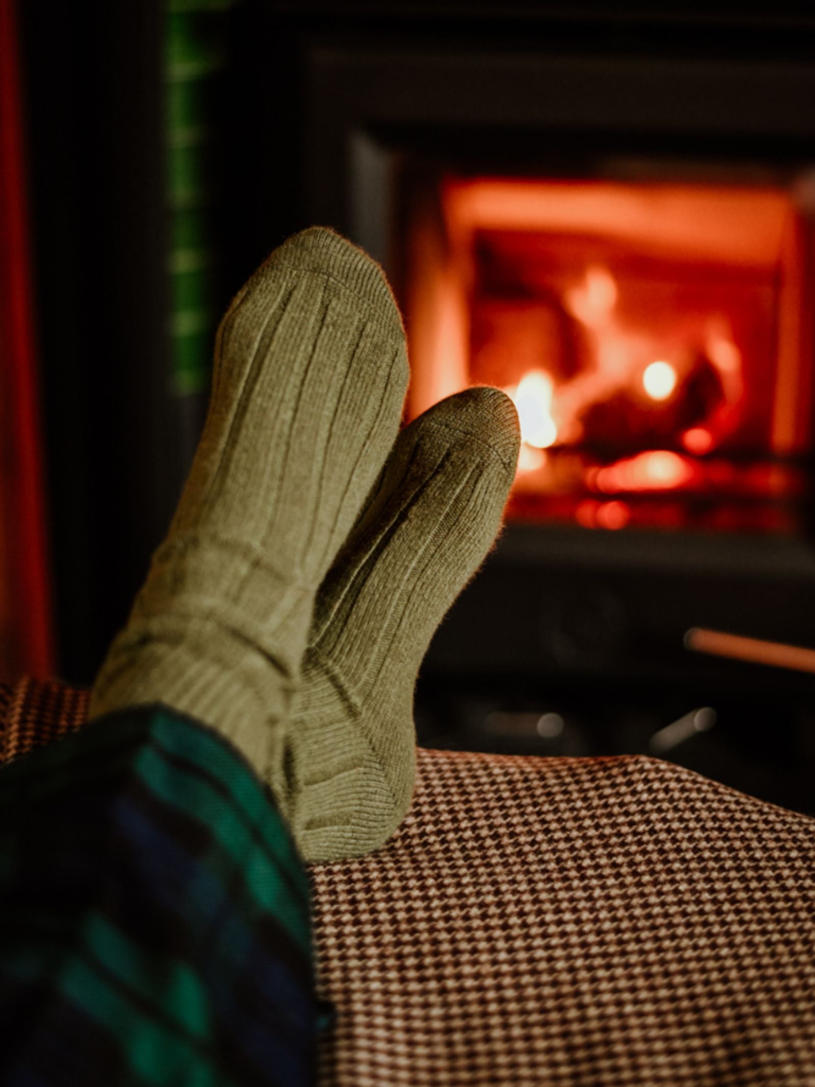 Someone rests their feet, clad in Campbell's of Beauly Rib Cashmere Socks and plaid pajama pants, on a patterned surface by a glowing fireplace, creating a cozy and warm atmosphere.