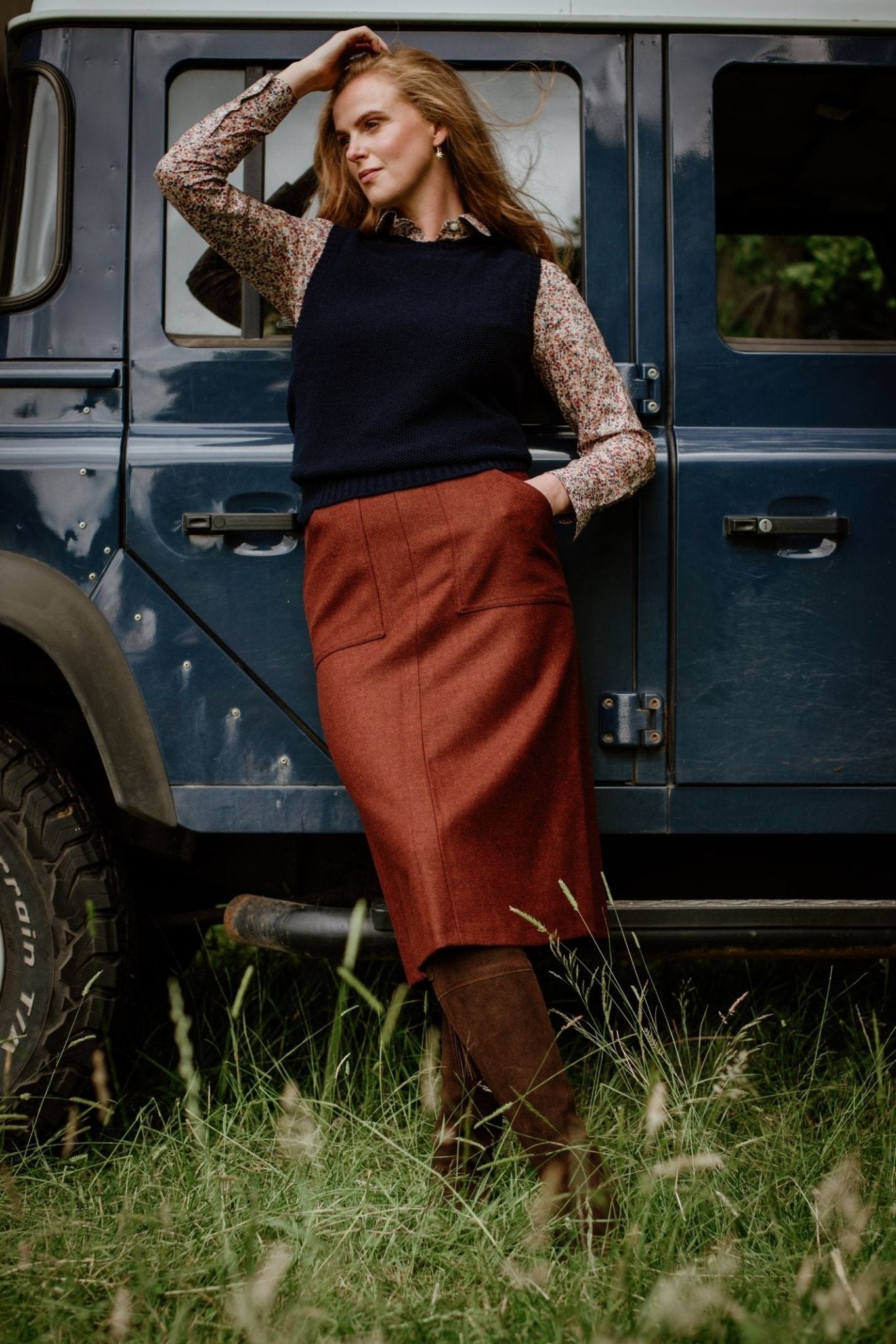 A woman stands in tall grass beside a dark blue off-road vehicle, wearing a floral blouse, navy sweater vest, Campbell's of Beauly A-Line Skirt, and brown boots, with one hand on her head and the other on the vehicle.