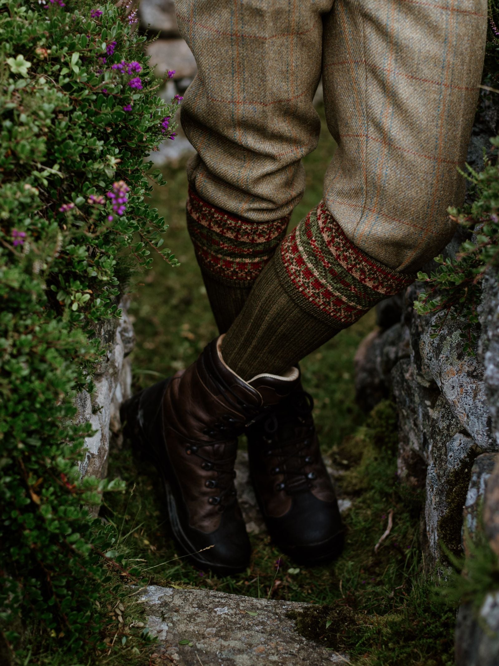 A person stands outdoors on a grassy path, wearing brown leather hiking boots and Campbells of Beauly Fairisle Shooting Socks made from a merino and acrylic blend. Green foliage, stone walls, and purple flowers line the path.