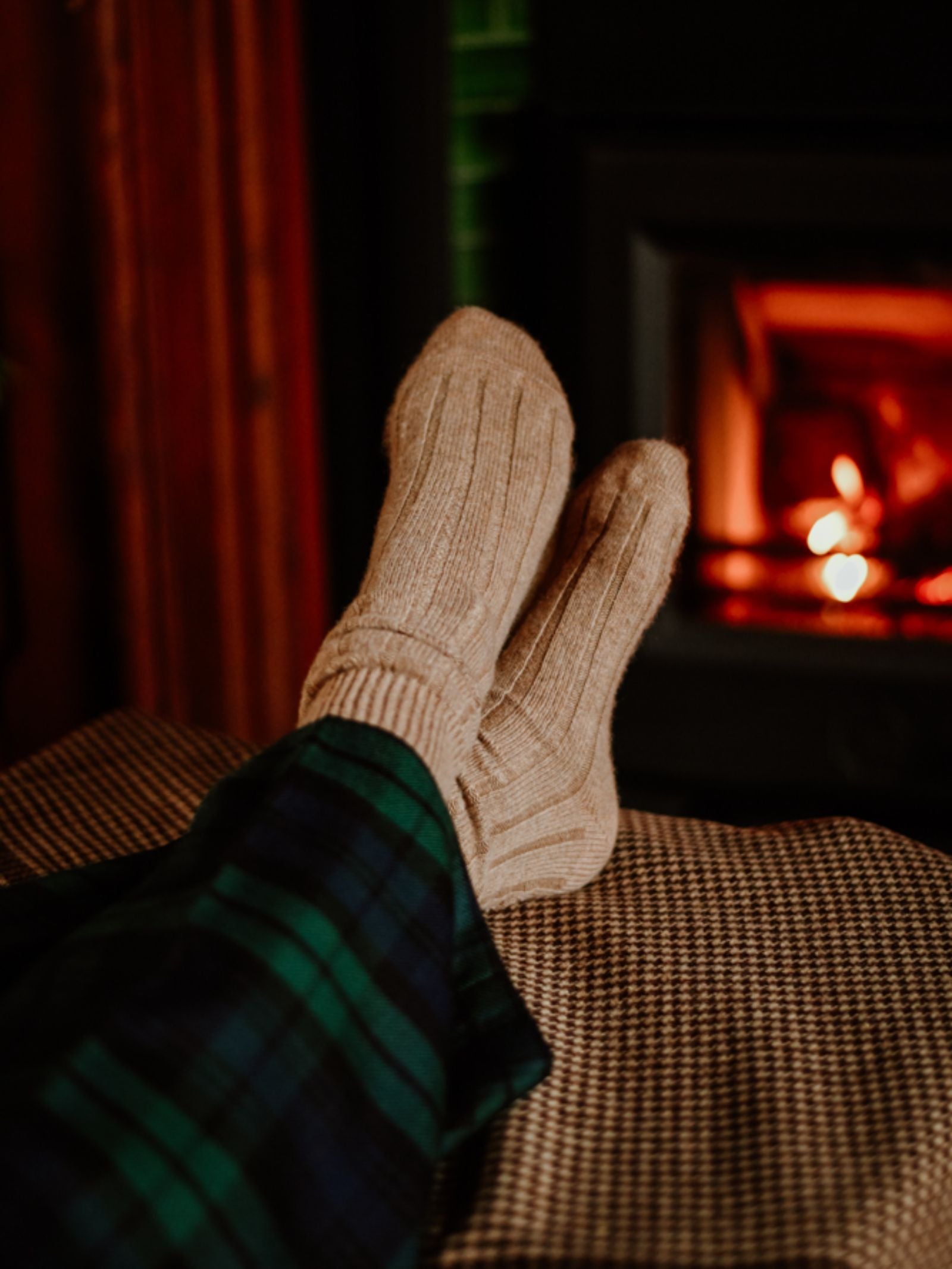 Wearing Campbell's of Beauly Rib Cashmere Socks and green plaid pajama pants, someone relaxes with their feet up by a warm fireplace, enjoying a cozy and comfortable atmosphere.