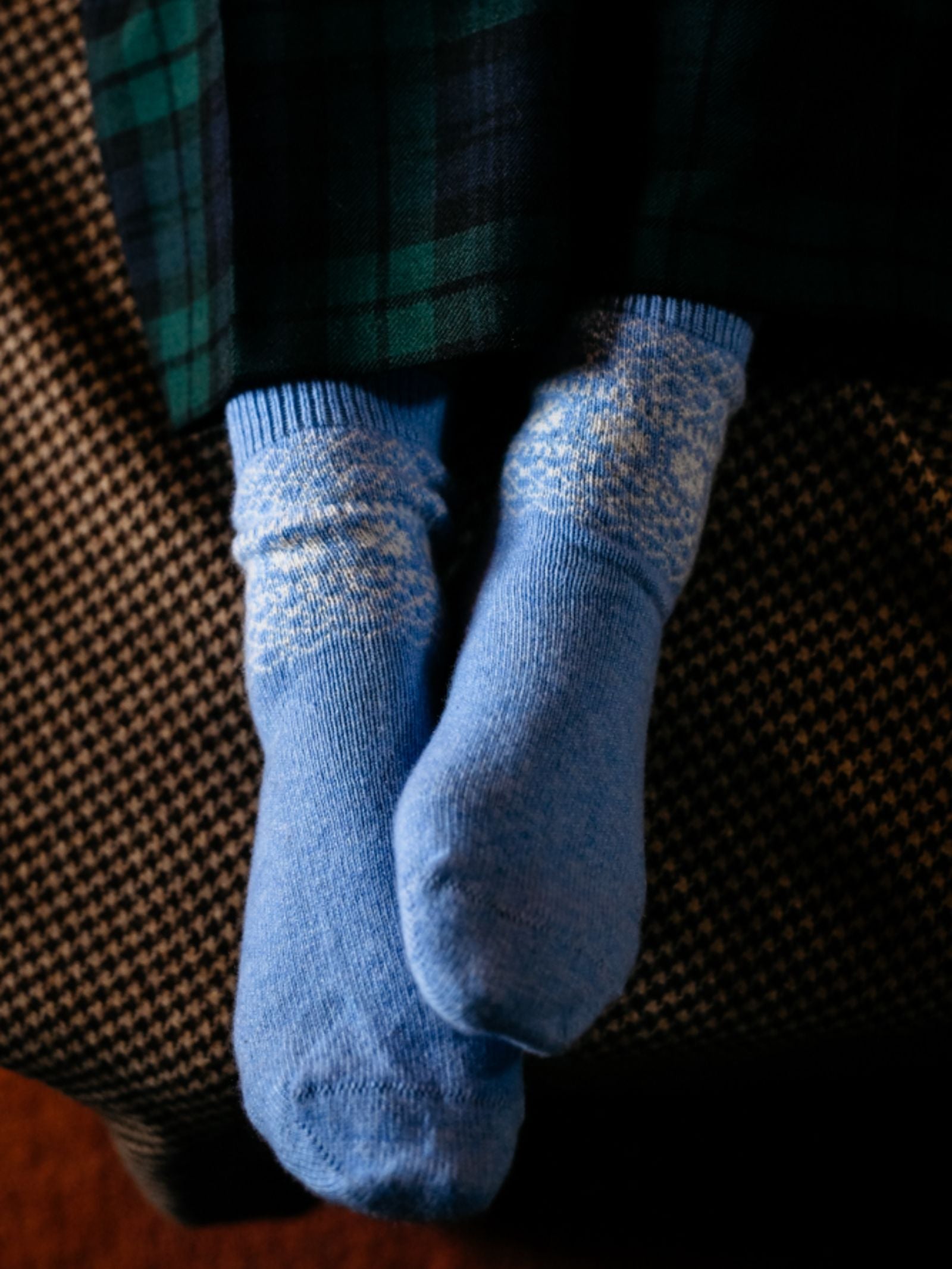 A person wearing Campbell's of Beauly Two-Colour Fairisle Cashmere Socks, featuring a white snowflake pattern, sits with their feet together on a textured brown surface, dark green and navy plaid pants visible.