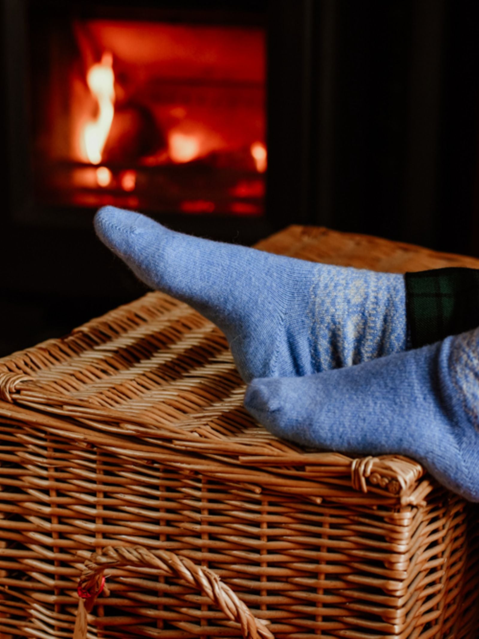 Two feet in Campbell's of Beauly Two-Colour Fairisle Cashmere Socks rest on a wicker basket before a glowing fireplace, creating a cozy, warm ambiance.