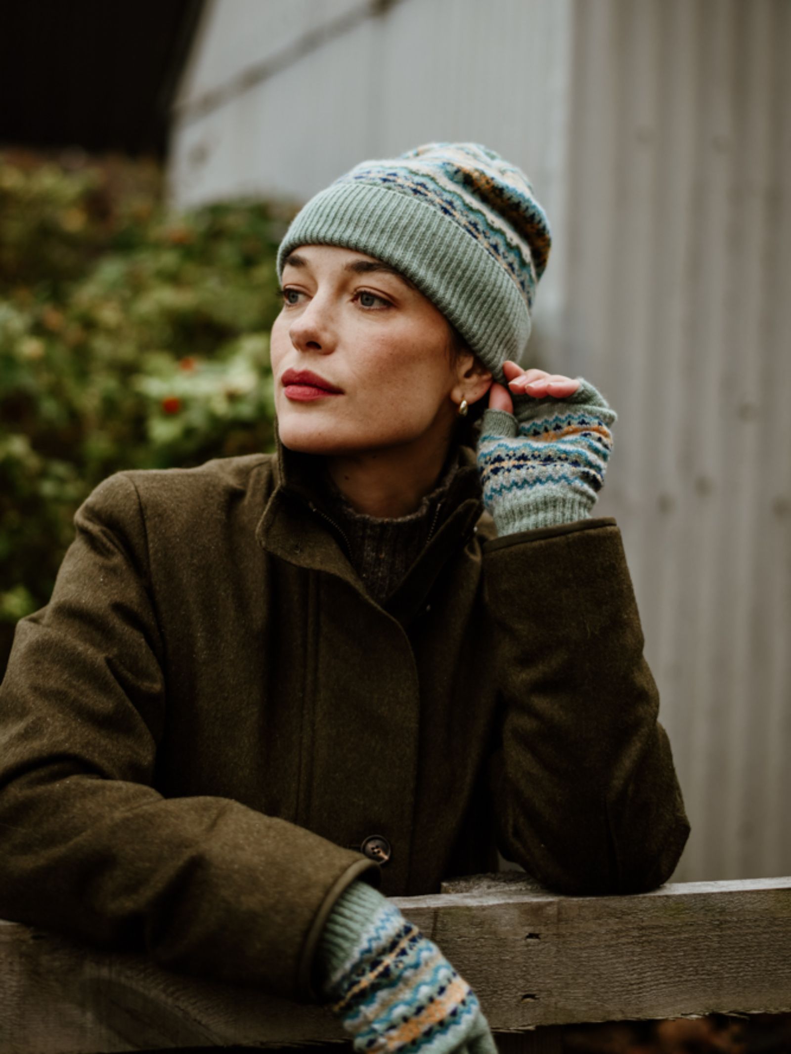A woman in the Campbell's of Beauly Lambswool Fairisle Hat leans on a wooden fence, wearing an olive coat and matching gloves. She gazes thoughtfully away, with greenery and a metal building behind her—a true winter essential.
