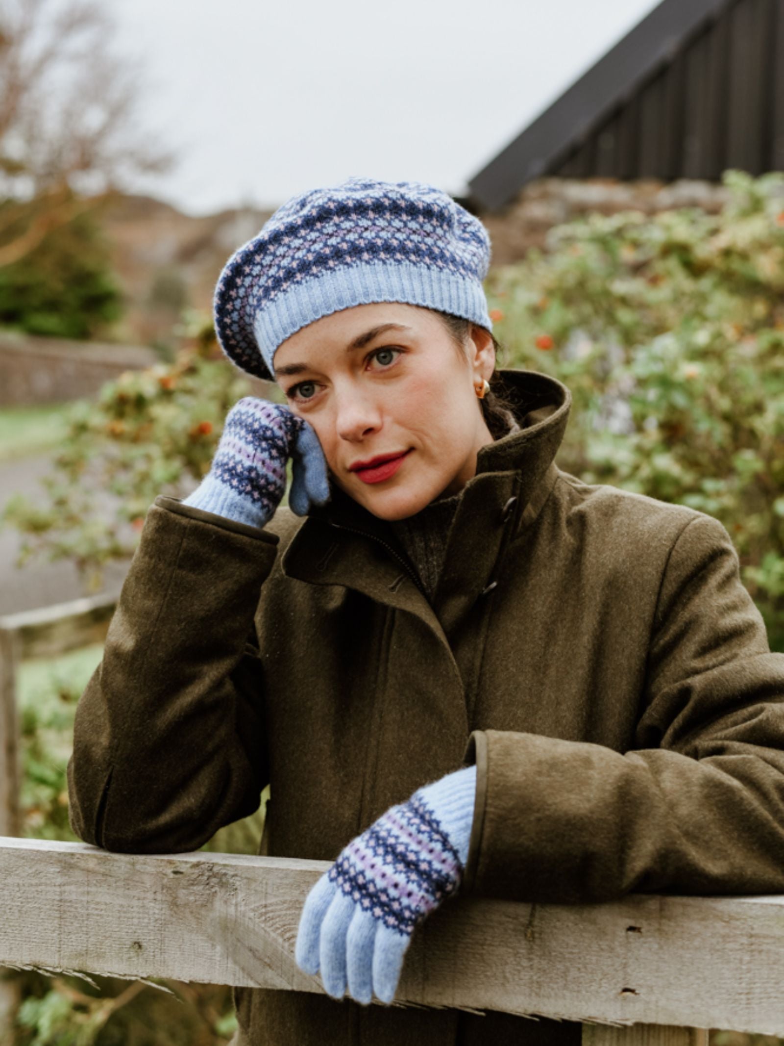 A woman wears the Campbell's of Beauly Fairisle Beret with matching gloves and a dark coat, leaning on a wooden fence outdoors. She looks thoughtful amid green foliage and a blurred building in the background.
