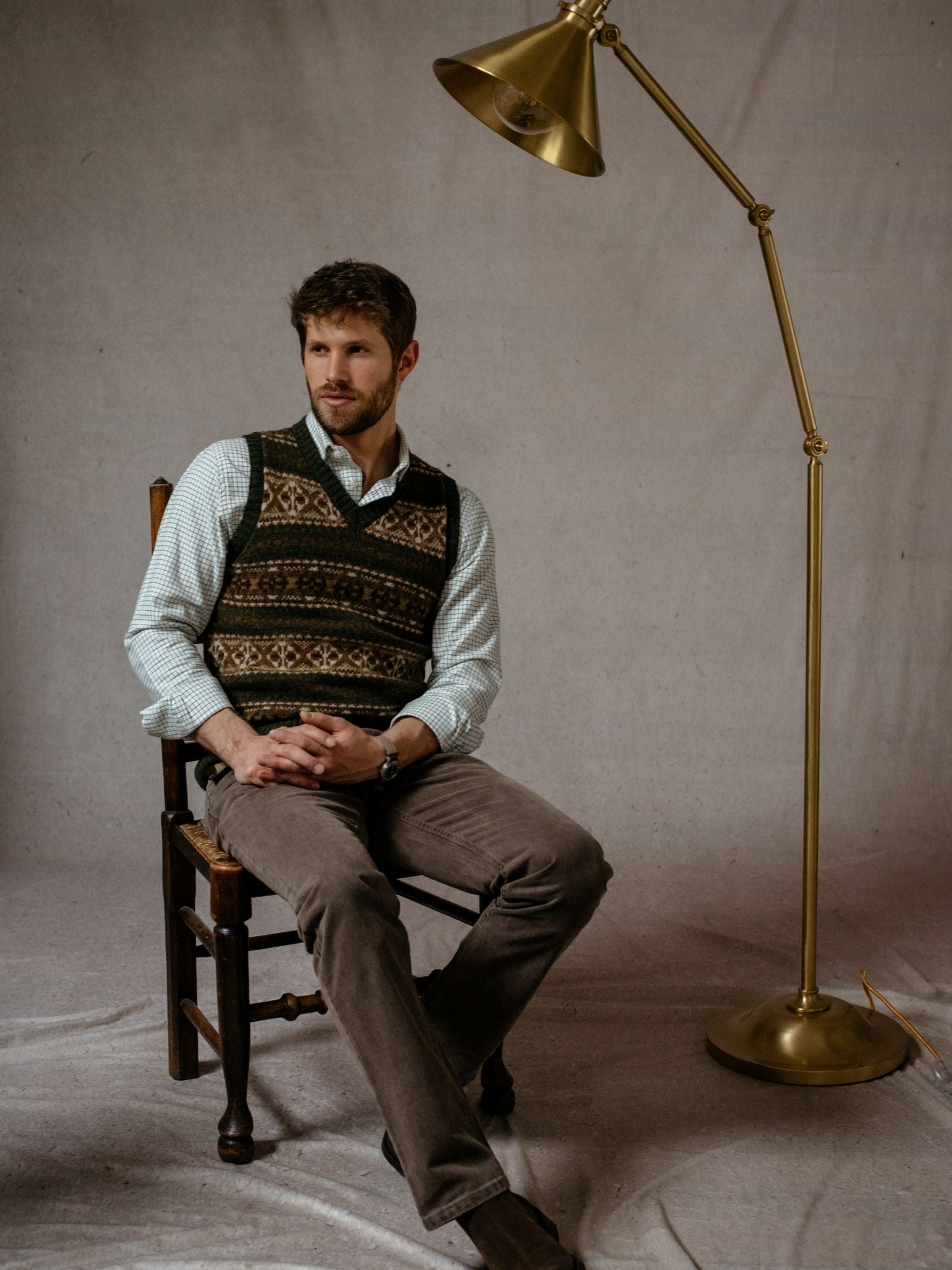 A man with short brown hair and a beard sits cross-legged on a wooden chair wearing the Campbells of Beauly Fairisle Slipover over a light shirt and brown pants. A tall gold floor lamp is beside him; the backdrop is plain neutral fabric.