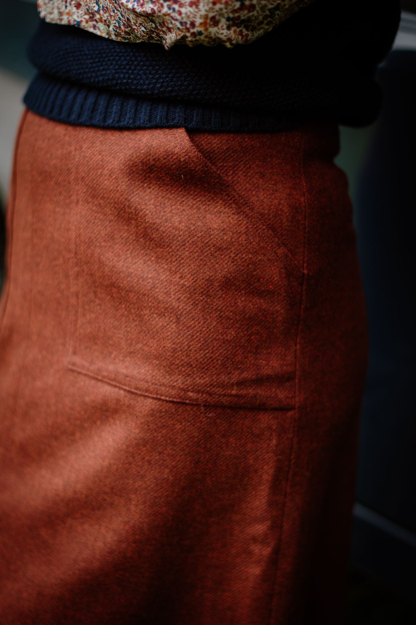 A close-up shows a person wearing Campbell's of Beauly A-Line Skirt in rust-colored tweed, paired with a dark knit sweater and a printed blouse. Only the torso and part of the skirt are visible.