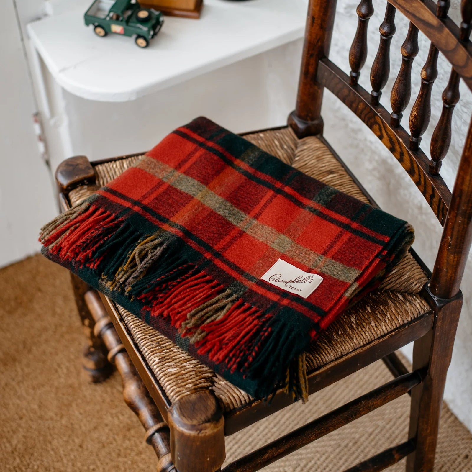 A Campbell's of Beauly Lambswool Blanket in Dark Maple Tartan is folded on a wooden spindle chair. On the windowsill behind are a green toy truck, green glass bottle, and small brown radio—classic Yorkshire charm accents.