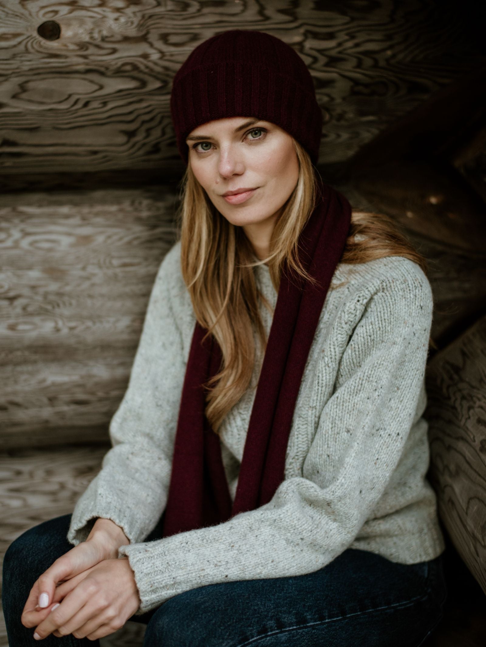 A woman with long blonde hair wears a maroon Campbells of Beauly Recycled Cashmere & Merino Scarf and beanie, paired with a light gray sweater and dark jeans, sitting calmly against a rustic wooden wall—a true winter essential.