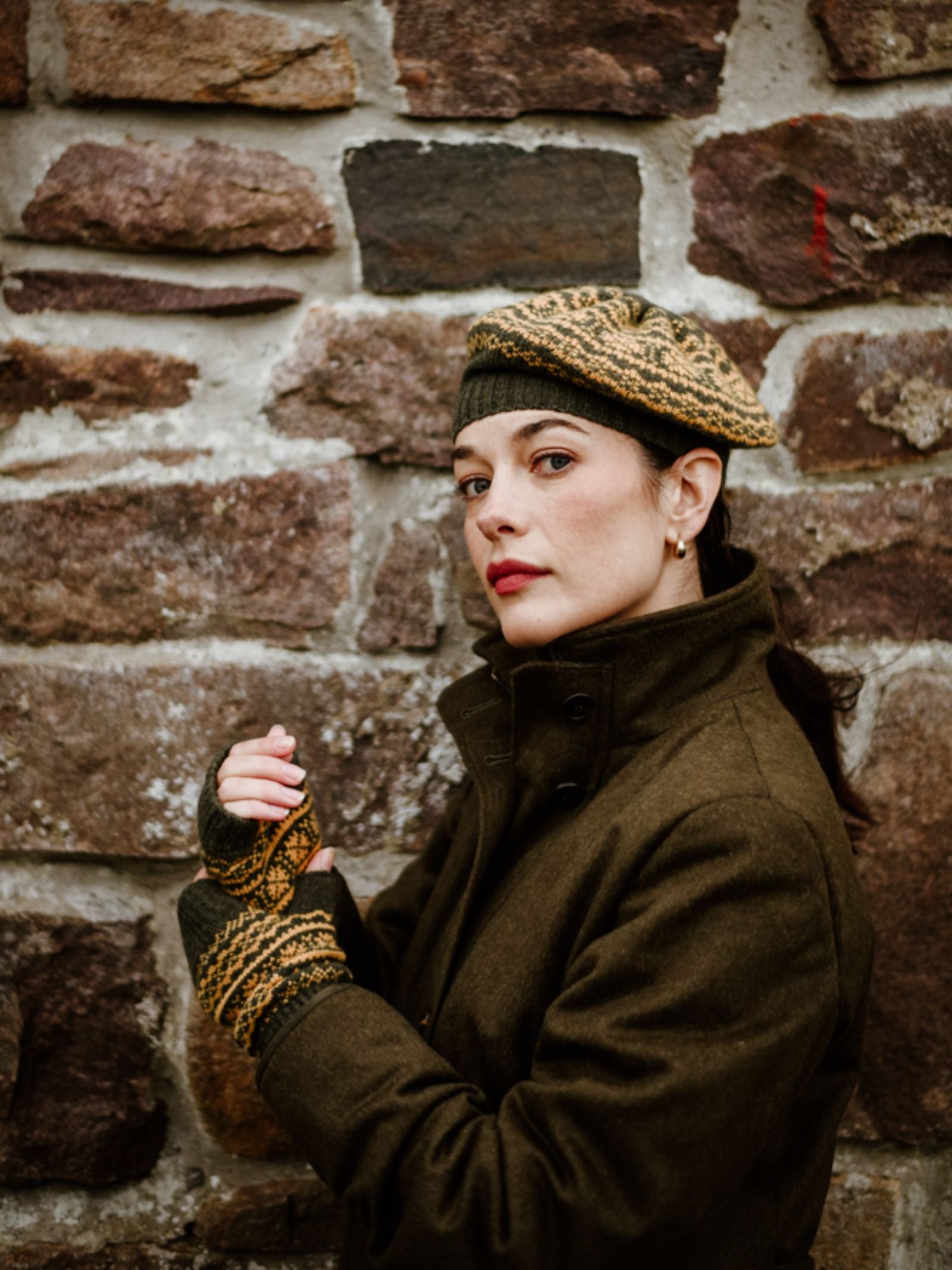 A woman in a dark coat and Campbell's of Beauly Two-Colour Fairisle Beret stands before a stone wall, glancing over her shoulder; the Scottish knitwear lends a classic charm to the scene.