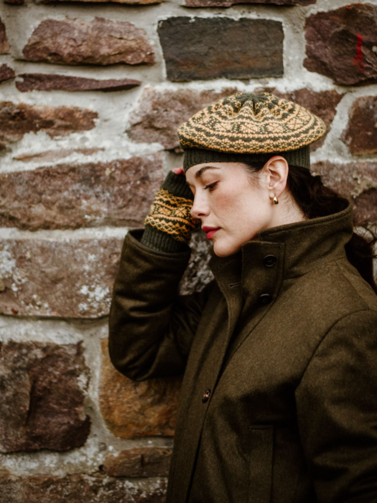 A woman wears a Two-Colour Fairisle Beret by Campbell's of Beauly with matching lambswool fingerless gloves and a green coat, standing against a stone wall with her hand to her head and eyes closed, looking thoughtful.