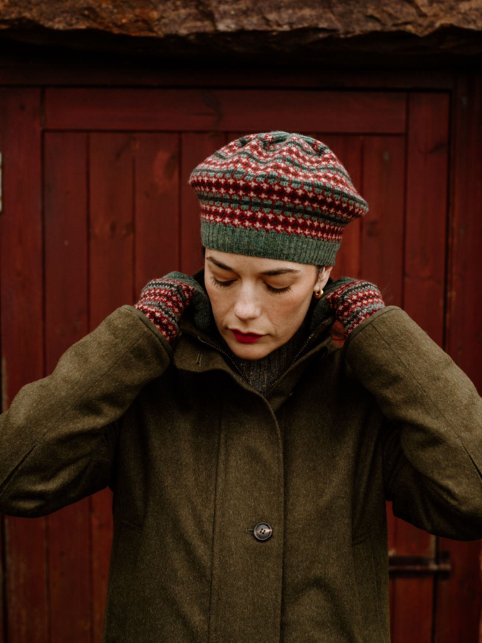 A person in an olive green coat adjusts their collar while wearing the Fairisle Beret by Campbell's of Beauly, standing in front of a weathered red wooden door.