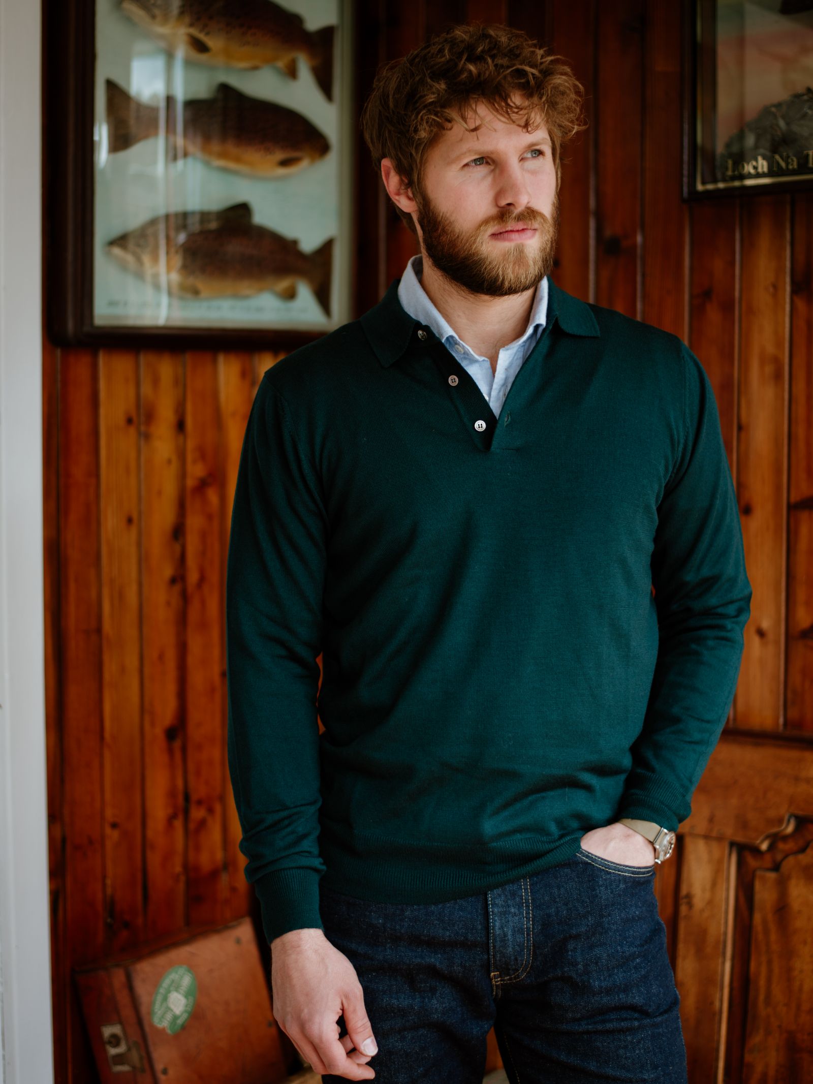 A man with curly brown hair and a beard stands indoors against wooden paneling, wearing Campbells of Beauly’s Merino Shirt Jumper in forest green over a blue collared shirt and dark jeans. One hand is in his pocket; framed fish illustrations hang behind him.