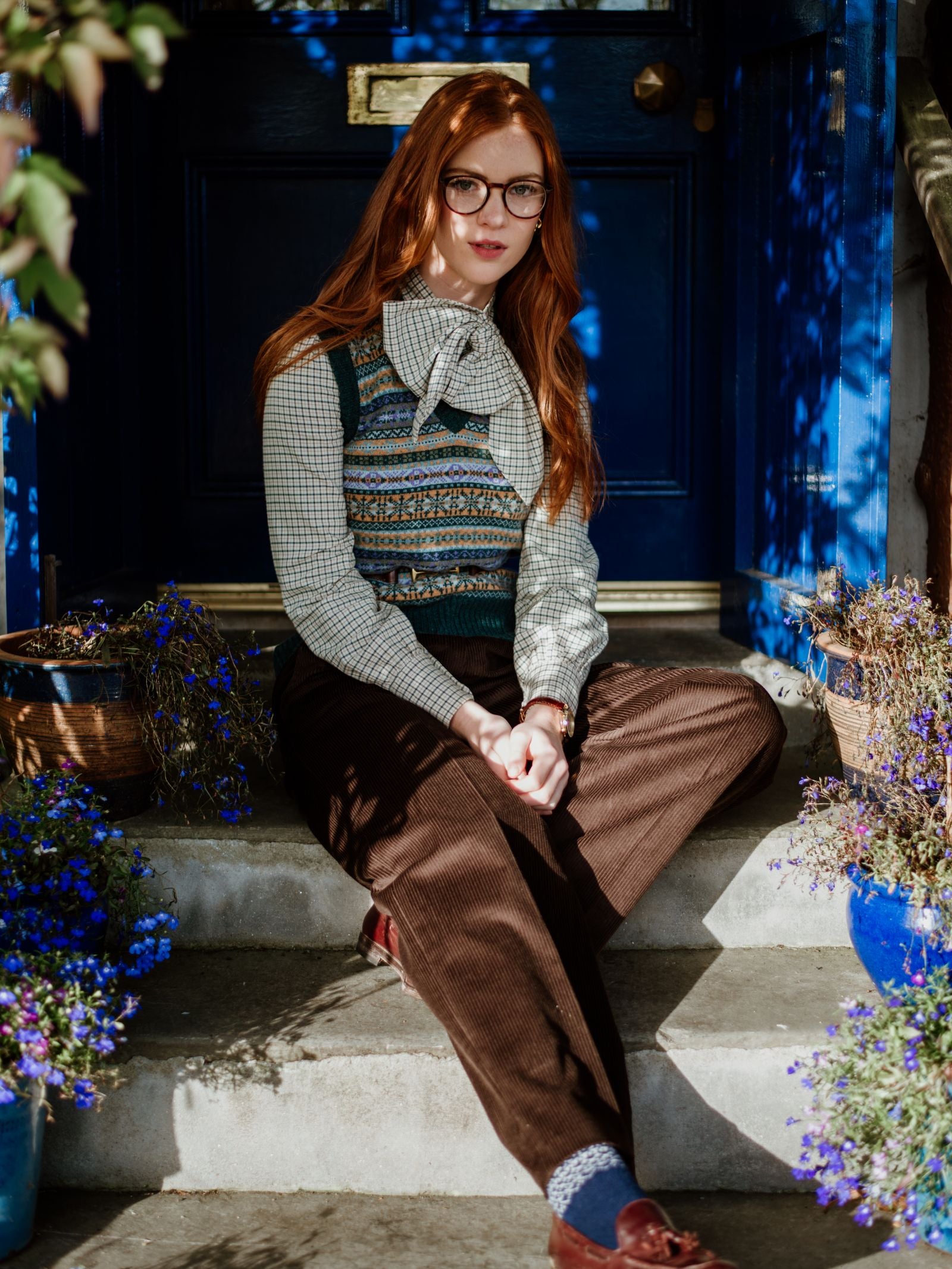 A young woman with long red hair and glasses sits on stone steps by a blue door amid blue and purple flowers, wearing a patterned vest, checked blouse with bow, brown corduroy pants, and Campbell’s of Beauly Two-Colour Fairisle Cashmere Socks.