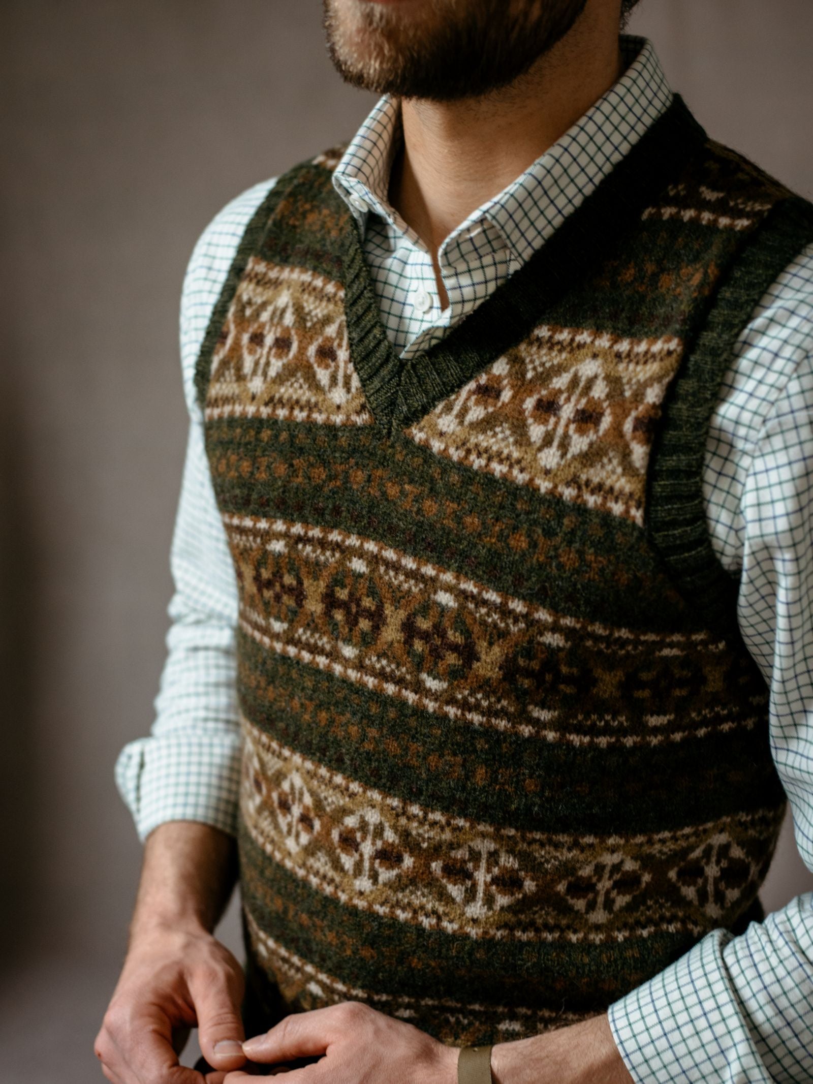 A bearded man wears the Campbells of Beauly Fairisle Slipover, a sleeveless Shetland wool sweater vest with a Fair Isle pattern, layered over a white checkered button-down, standing with hands clasped against a softly blurred background.