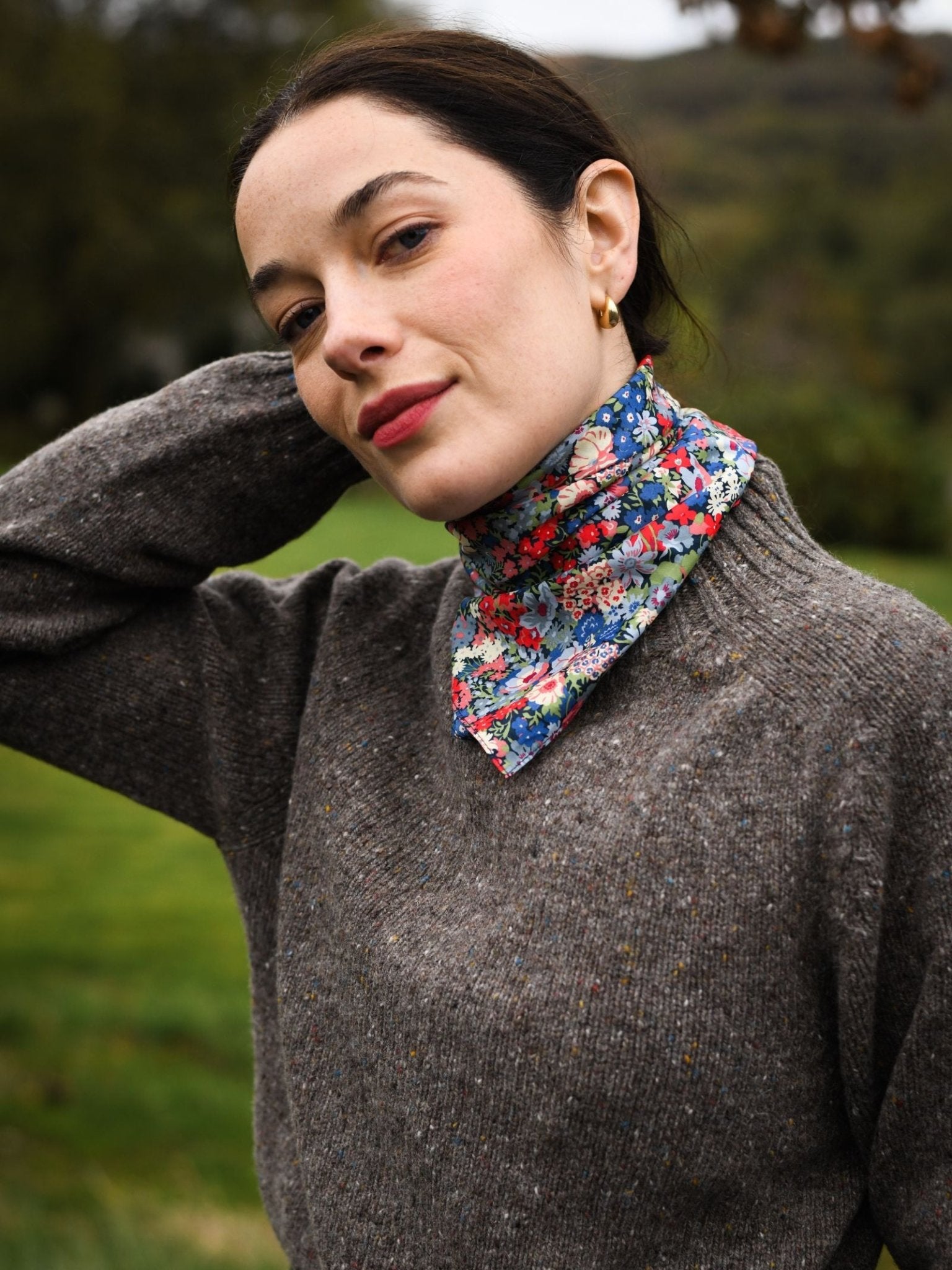 A woman with fair skin and dark hair poses outdoors, wearing a Campbell's of Beauly Square Headscarf in a colorful floral print around her neck and a gray knit sweater, with greenery softly blurred in the background.