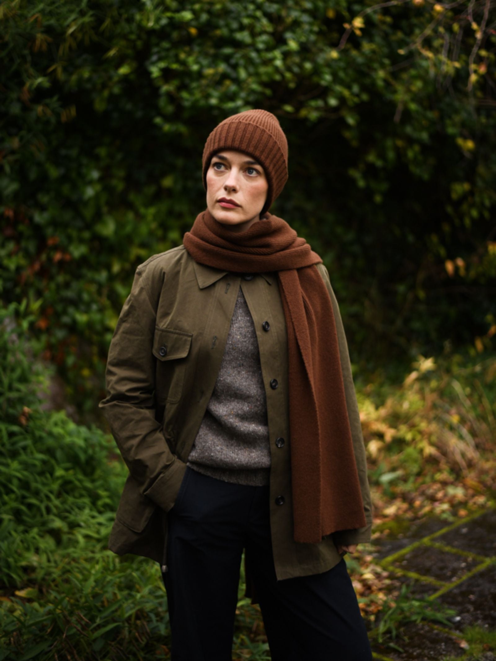 A woman stands outdoors before green foliage, wearing Campbell's of Beauly Rib Hat in brown—an essential winter accessory—with a matching scarf, olive jacket, grey sweater, and dark pants. She gazes thoughtfully to the side with one hand in her pocket.