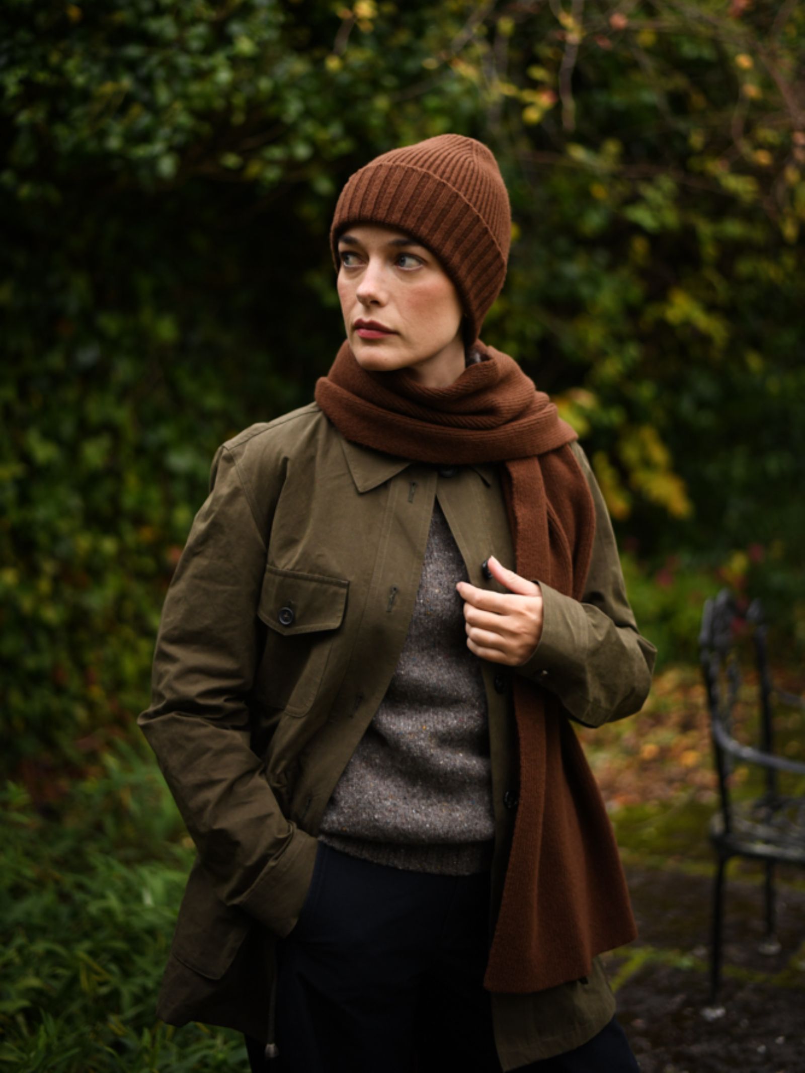 A person wears the Campbell's of Beauly Rib Hat—knitted in the UK from brown wool—paired with a brown scarf, olive green jacket, and gray sweater, standing outdoors before green foliage and looking to the side with one hand in their pocket.