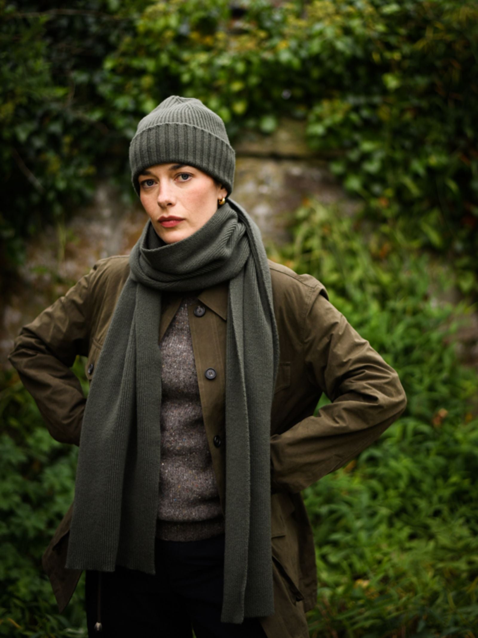 A woman wearing the Campbell's of Beauly Rib Hat—a green wool winter essential made in the UK—pairs it with a matching scarf and brown coat, standing outdoors among lush greenery with a neutral expression and hands on hips.