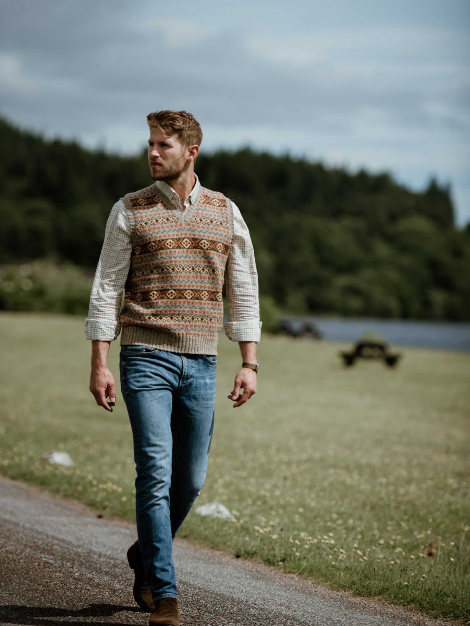 A man with short brown hair and a beard walks by a lake in blue jeans, brown shoes, and a Campbells of Beauly Windowpane Shirt under a patterned knit vest. Green trees and cloudy sky set a cool, peaceful background.