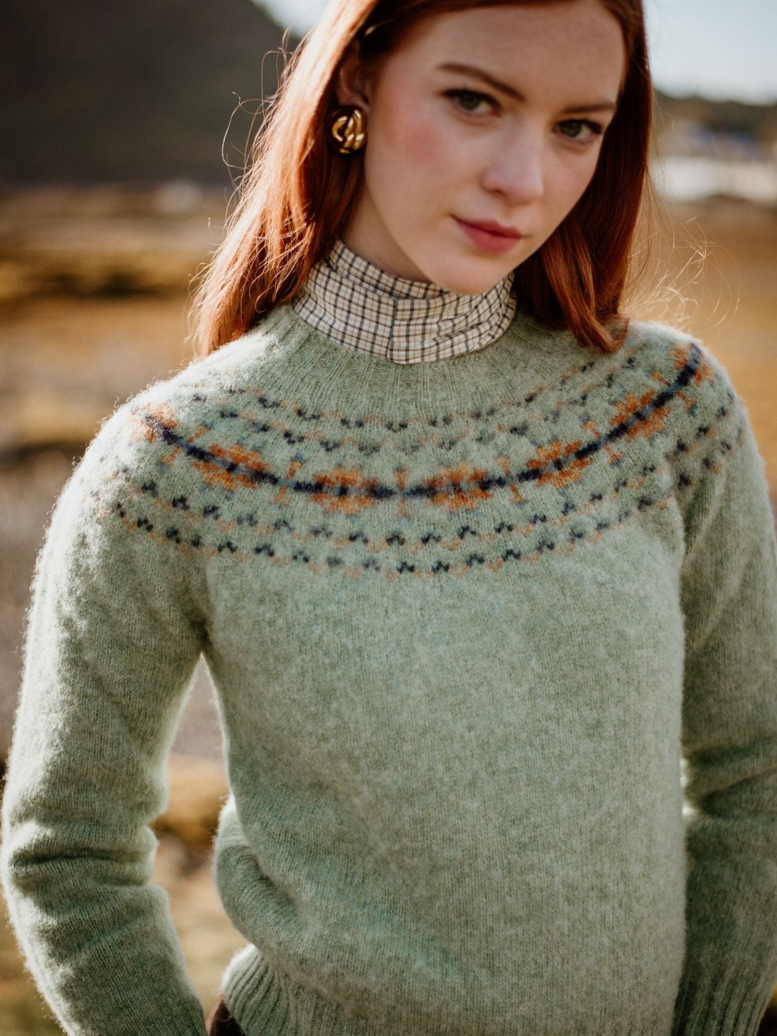A woman with long red hair wears the Campbell's of Beauly Cropped Fairisle Crew Jumper, featuring a colorful patterned yoke, over a checked shirt while standing outdoors against a blurred natural background.