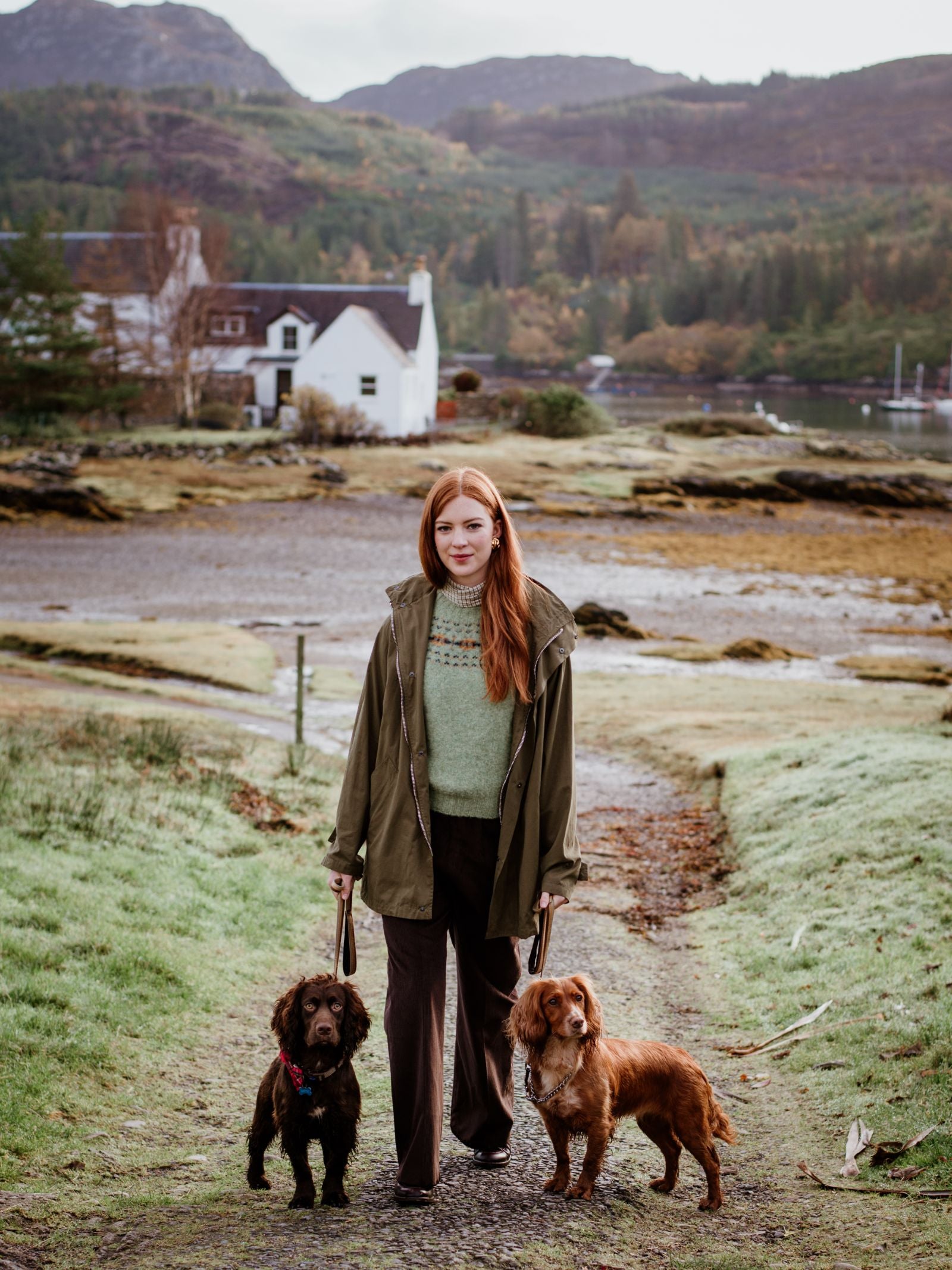 A woman with long red hair stands on a grassy path holding two brown dogs. She wears the Mont Coat by Campbells of Beauly—an olive outer layer for all seasons. Behind her: a white cottage, lake, and green hills under an overcast sky.