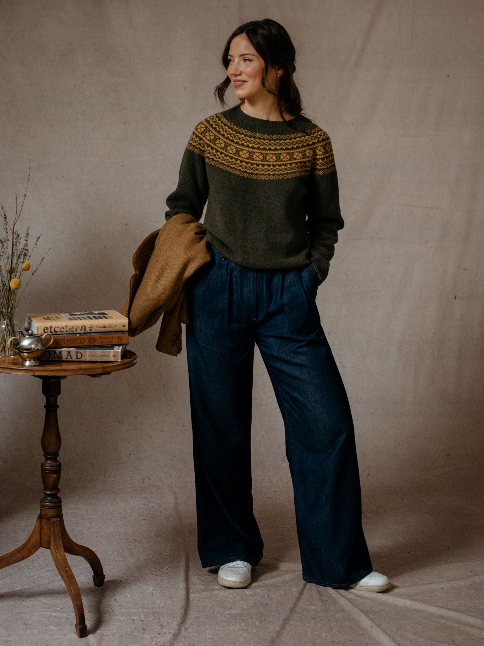 A woman stands indoors by a beige backdrop, wearing Campbells of Beauly Two-Colour Fairisle Yoke Jumper in dark green Geelong lambswool. She pairs it with loose blue jeans, white shoes, and holds a brown coat beside a table with books and a teapot.