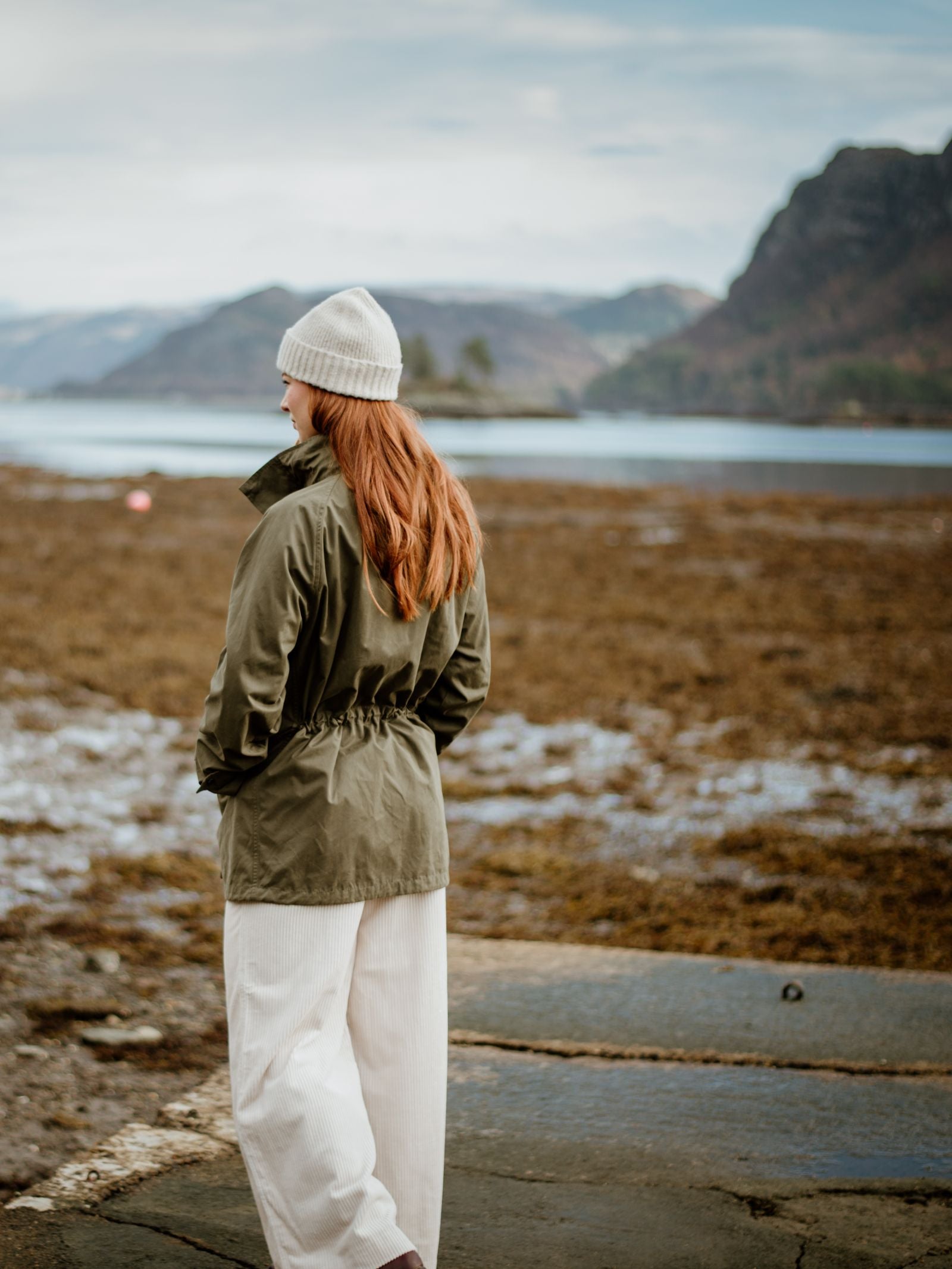 A woman with long red hair, wearing a white beanie and Campbells of Beauly Mont Coat in olive green, stands with her back to the camera on a rocky shore, facing distant mountains under a cloudy blue sky.