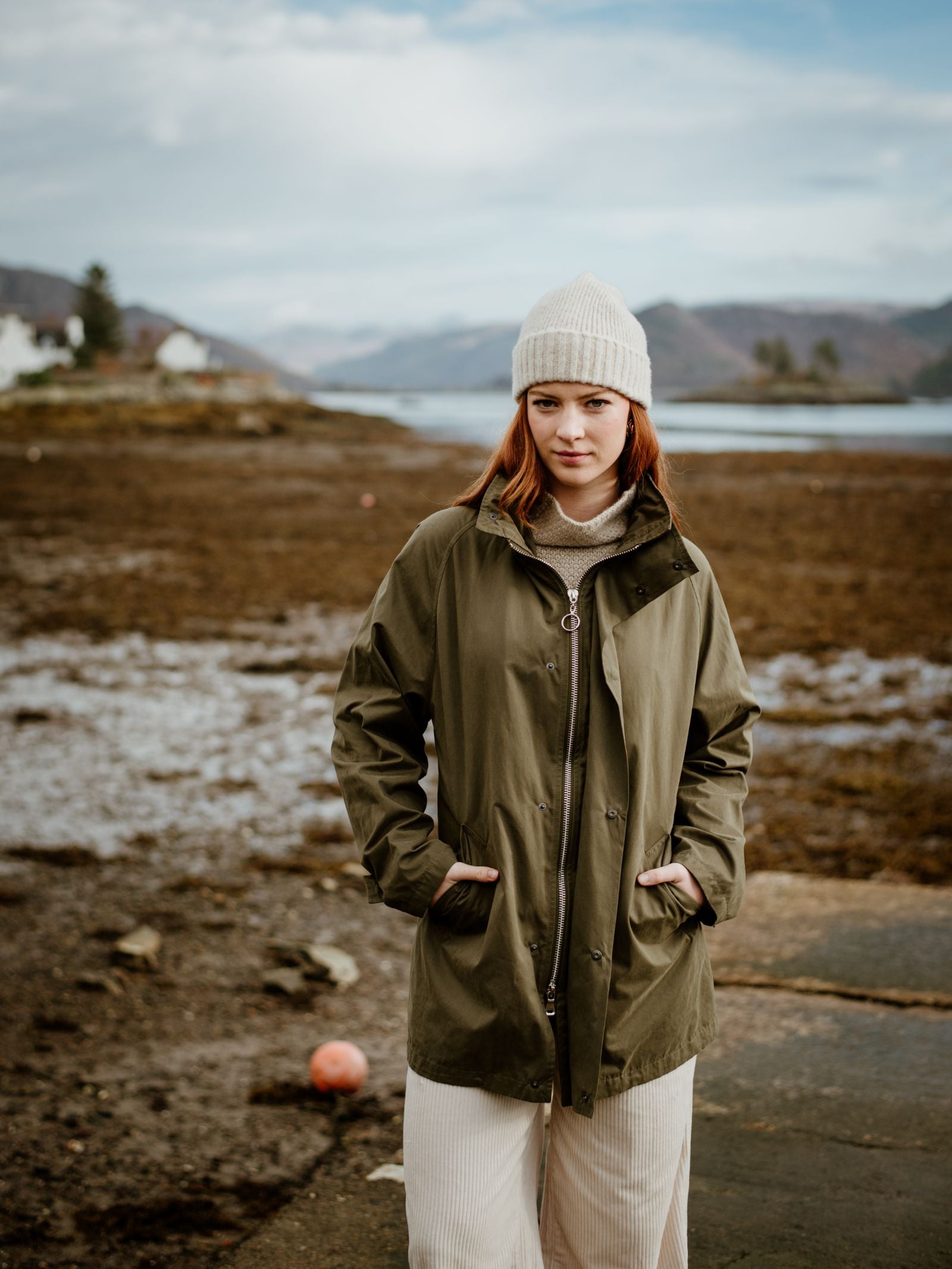 A young woman faces the camera outdoors on a rocky coast, hands in her pockets, wearing a Campbells of Beauly Lambswool Hat, an olive green coat, and cream pants under a partly cloudy winter sky.
