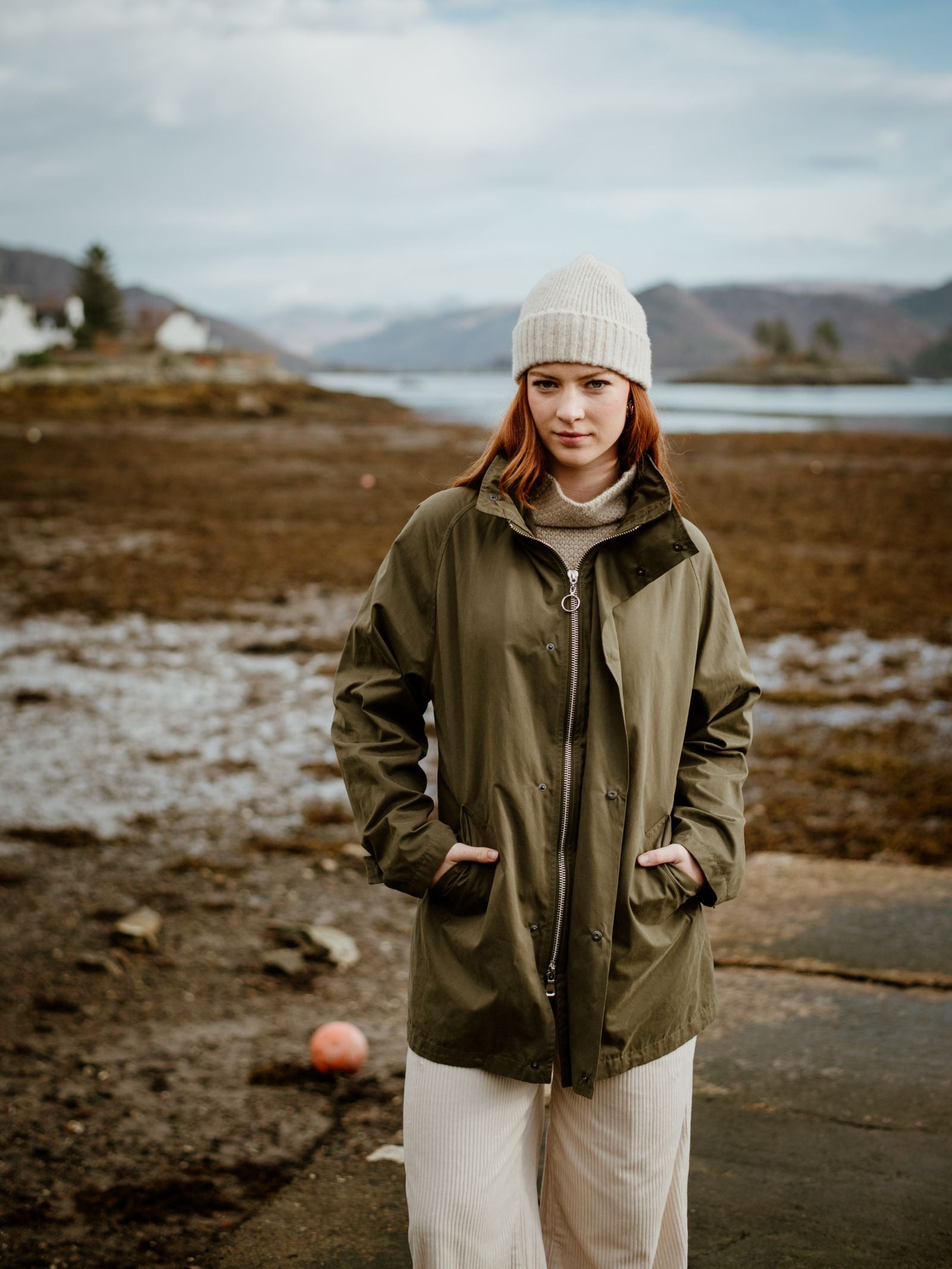 A young red-haired woman stands by a lake, hands in her pockets, wearing Campbells of Beauly Mont Coat in green, beige pants, and a cream knit beanie. Rocky terrain, water, and distant mountains appear under a cloudy sky.