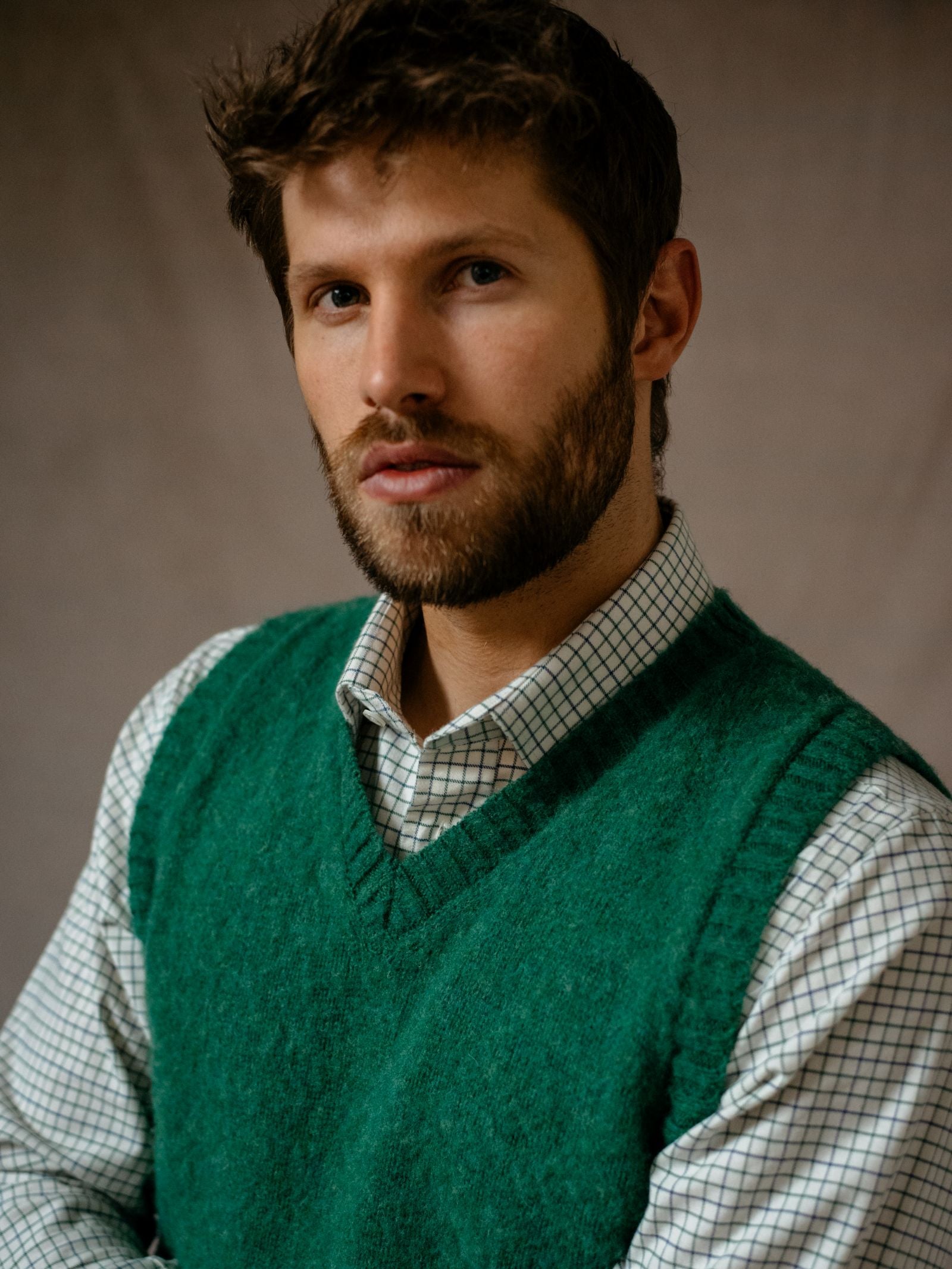 A man with short brown hair and a beard wears a green sweater vest over a white, checkered Campbells of Beauly Windowpane Shirt, facing forward with a neutral expression against a plain, softly lit background.