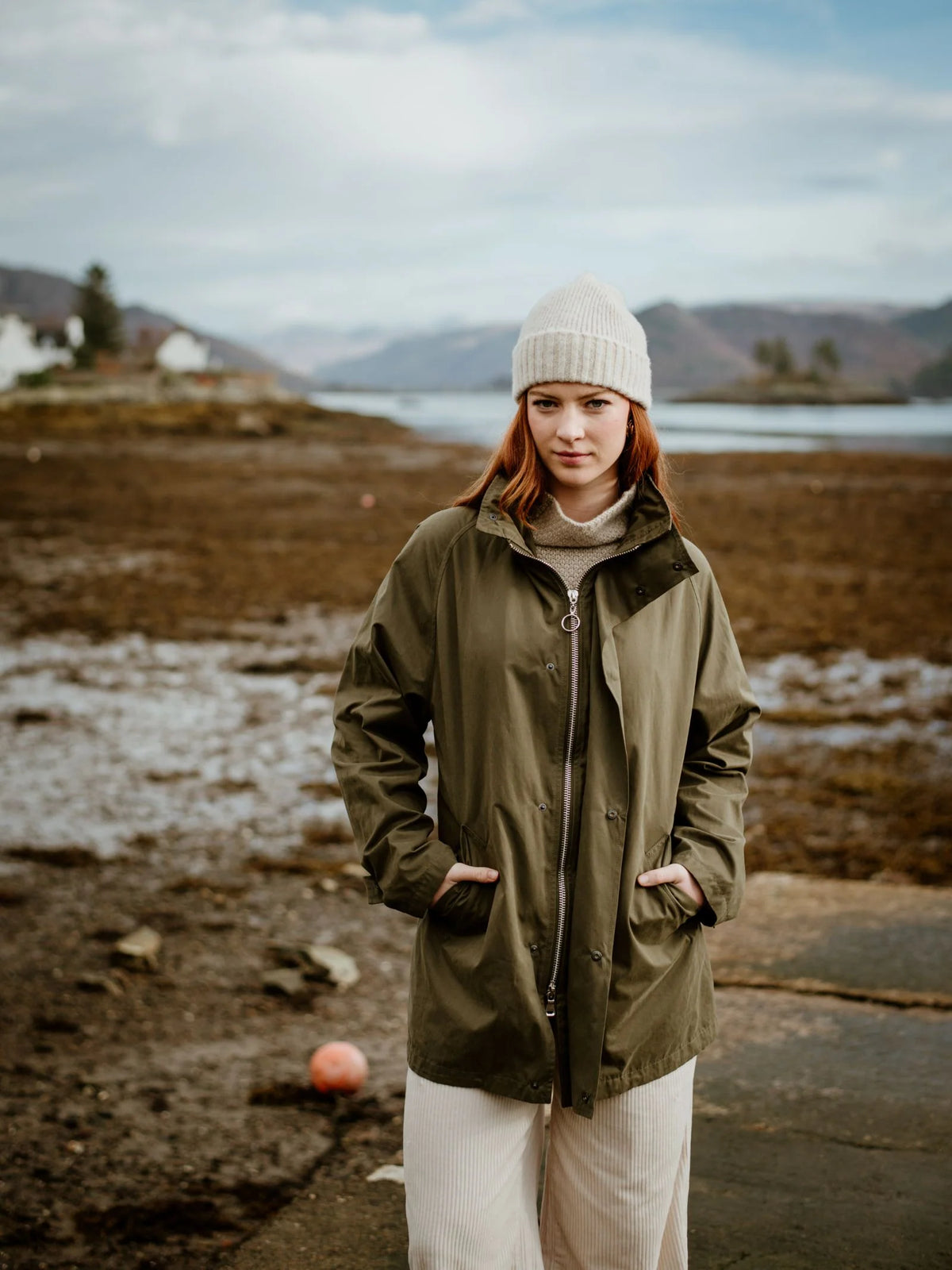 A person in a green jacket, light pants, and a Campbell’s of Beauly Lambswool Hat Cream stands outside by a rocky shore with mountains and water behind them.