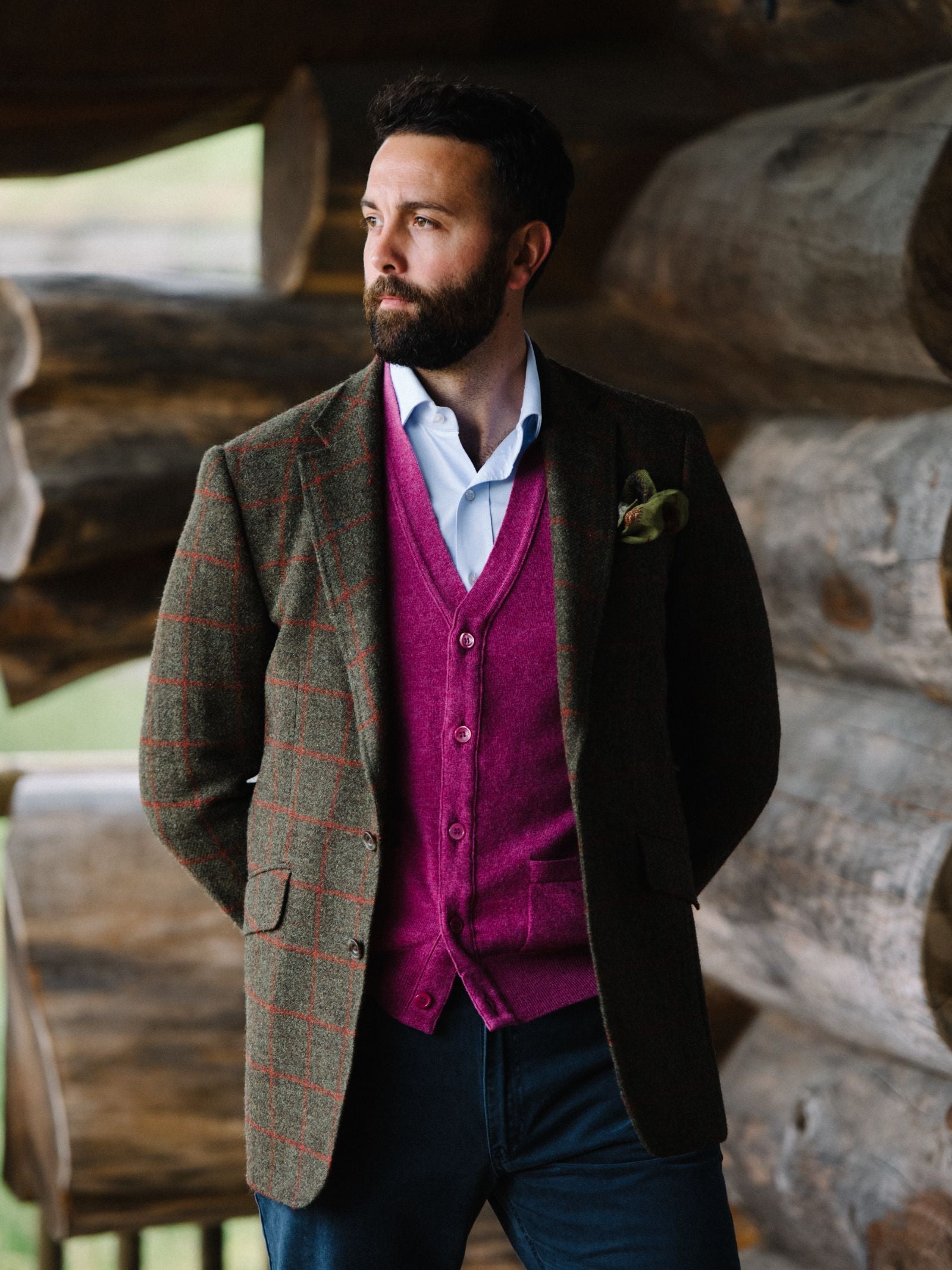 A bearded man stands before stacked logs, wearing a dark green plaid blazer, white shirt, dark jeans, and a bright magenta Geelong Lambswool Button Waistcoat by Campbells of Beauly. He gazes left with hands behind his back, looking thoughtful and stylish.