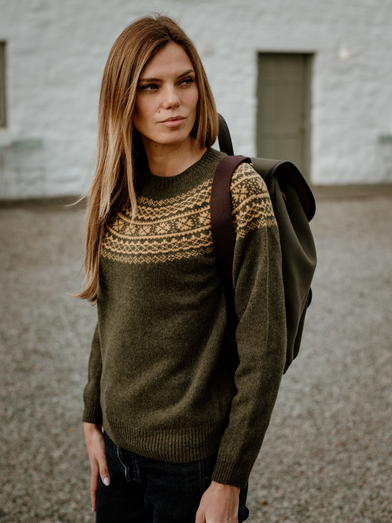 A woman with long brown hair wears the Two-Colour Fairisle Yoke Jumper by Campbells of Beauly, carrying a dark backpack on one shoulder. She stands outside on a gravel path before a white building with a green door and window.