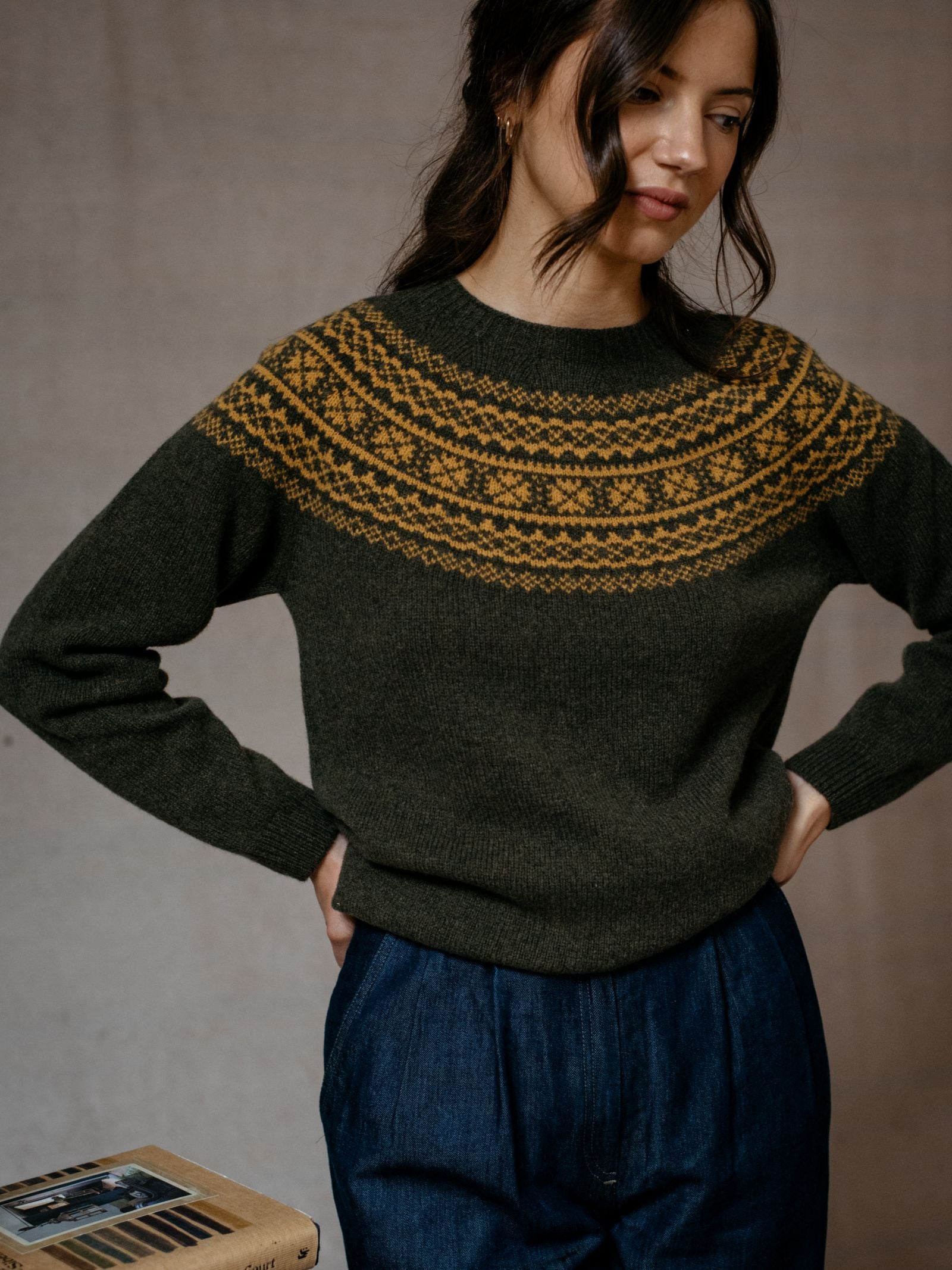 A woman with dark wavy hair wears Campbells of Beauly’s Two-Colour Fairisle Yoke Jumper in dark green Superfine Geelong lambswool with gold patterns and high-waisted blue pants, standing hands on hips beside a small table with photos.