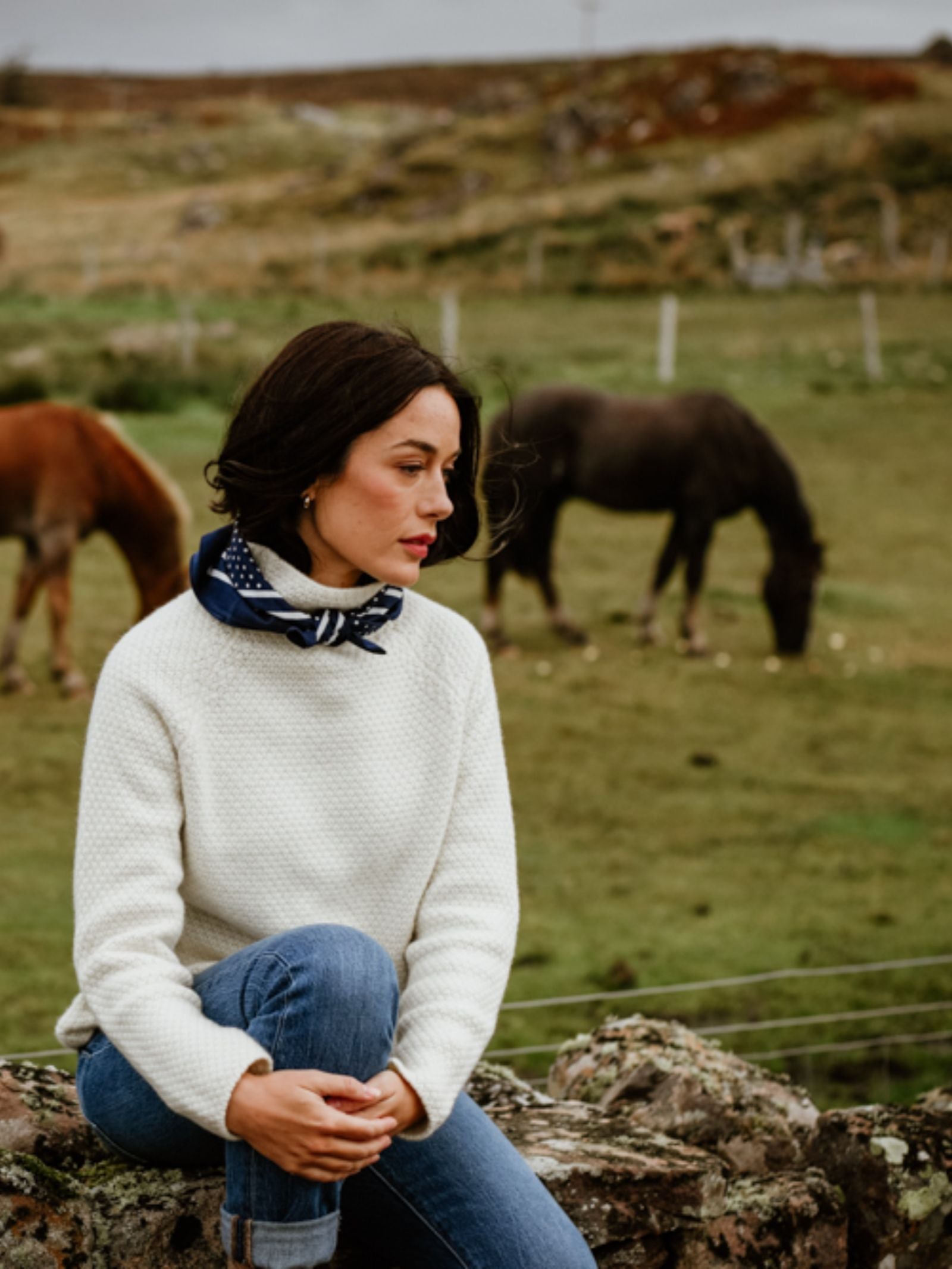 A woman with dark hair, wearing the Campbell's of Beauly Cashmere Funnel Neck Jumper and blue jeans, sits on a stone wall in a grassy field as two horses graze in the background.