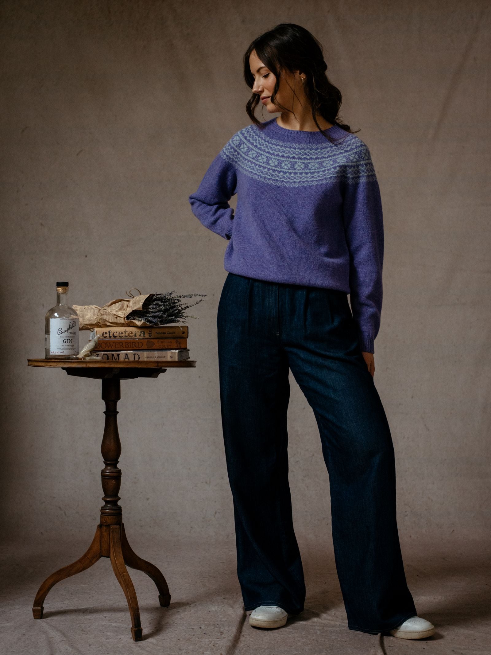 A woman with long dark hair stands indoors by a small round table, wearing Campbells of Beauly Two-Colour Fairisle Yoke Jumper in purple and wide-leg dark blue pants. The table holds a bottle, bread, herbs, and stacked books.