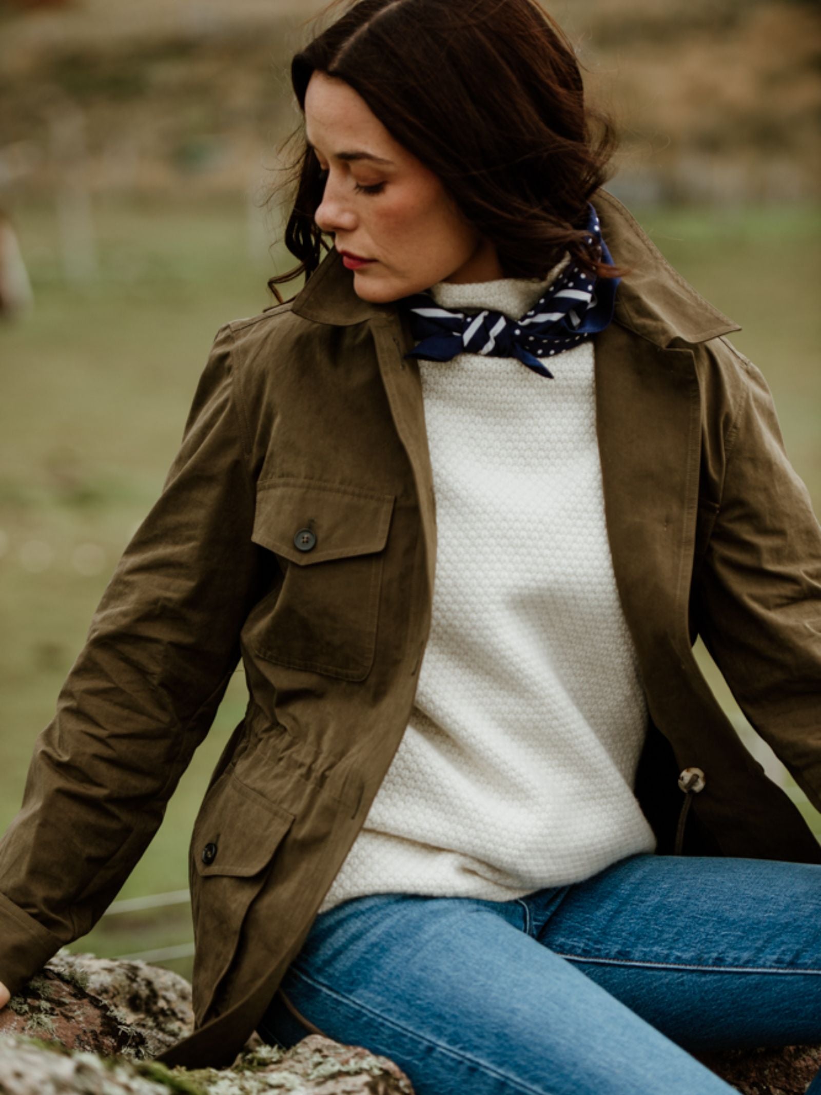 A woman with dark hair sits outdoors in a Campbell's of Beauly Auld Stock Aigas Field Jacket over a white textured sweater, blue jeans, and navy scarf, looking down to the side against a blurred natural background.