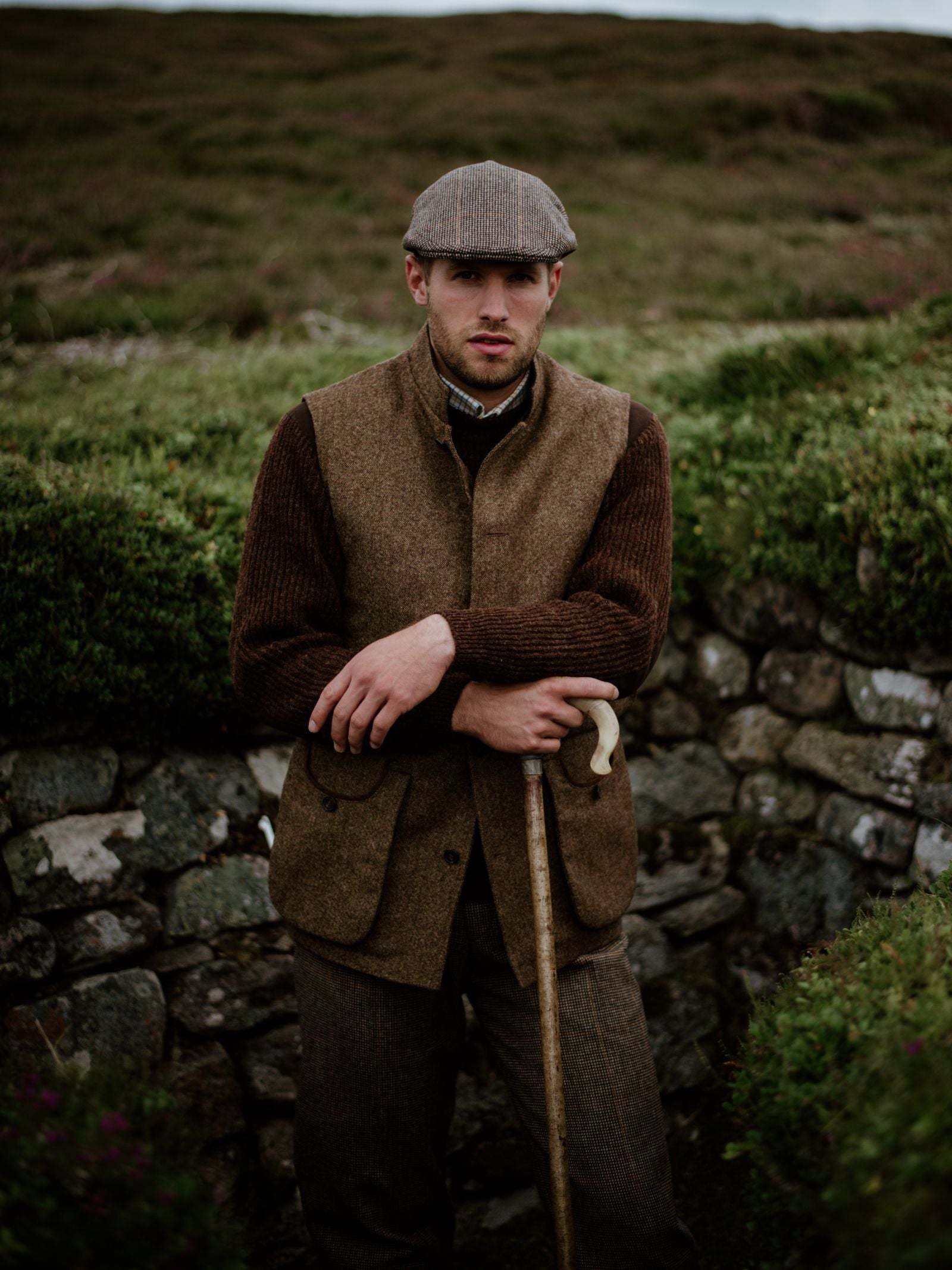 A man stands outdoors by a stone wall, wearing the Campbells of Beauly Field Nehru Gilet made from British moleskin with quilted lining. He holds a wooden walking stick against green shrubbery and moorland under an overcast sky.