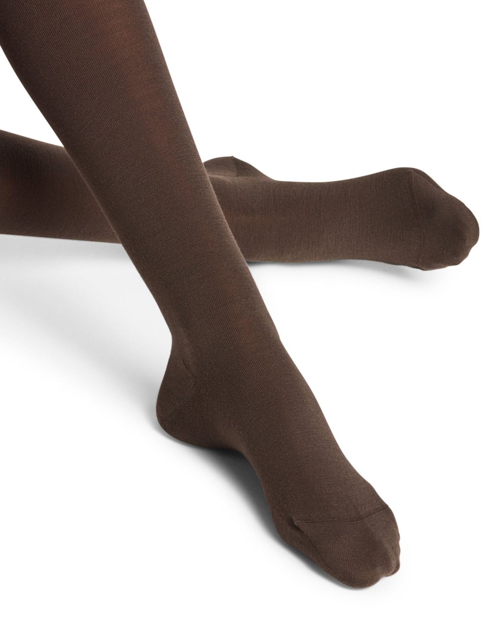 A person wearing FALKE Merino Tights in Dark Brown sits with one leg crossed over the other against a white background.