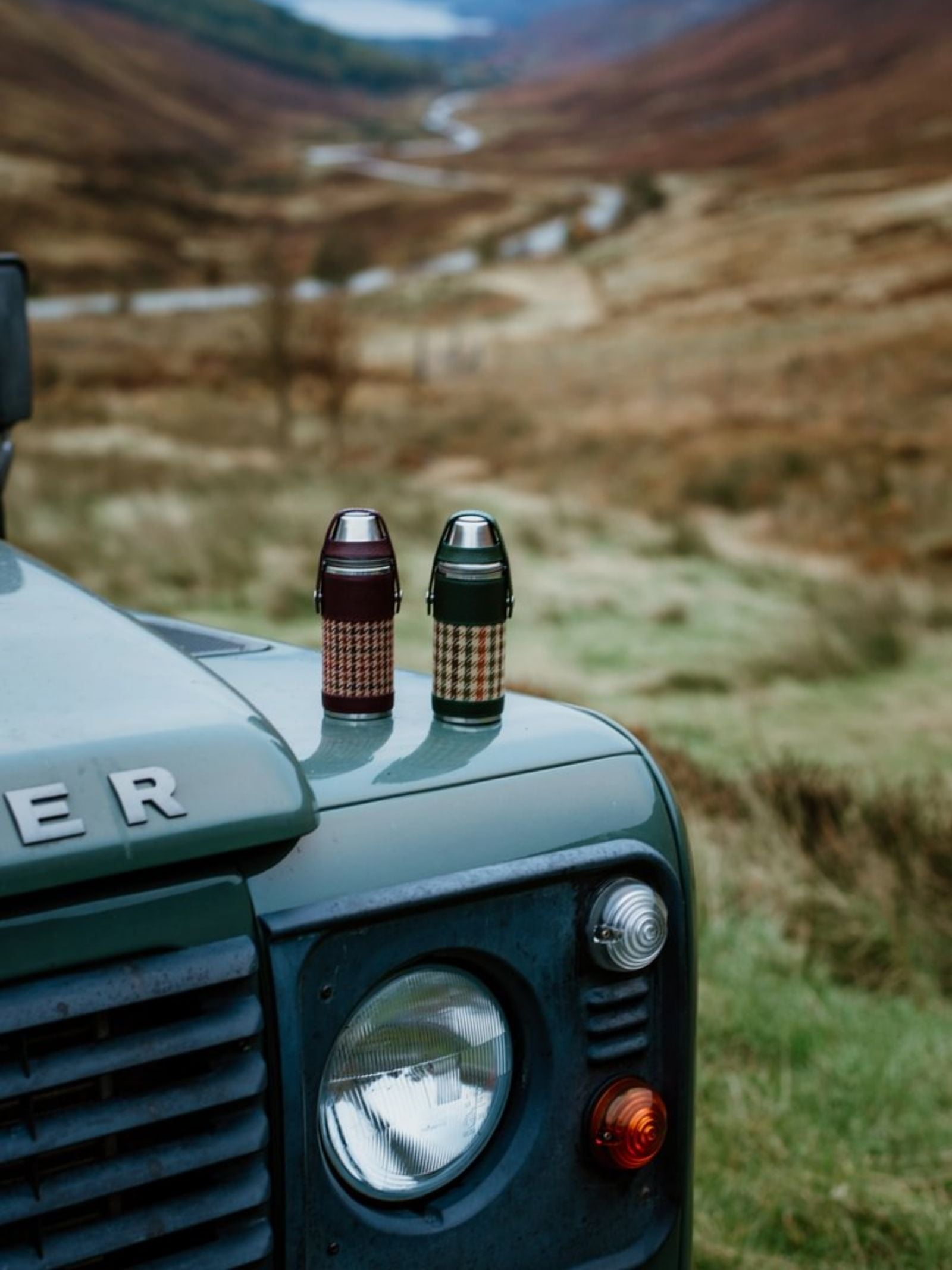 Two patterned thermos flasks, including the 8oz Bordeaux Hunter Flask by Campbell's of Beauly x Ettinger with Capra goat leather, rest on a green Land Rover’s hood in a grassy, hilly countryside beside a winding stream—celebrating British craftsmanship.